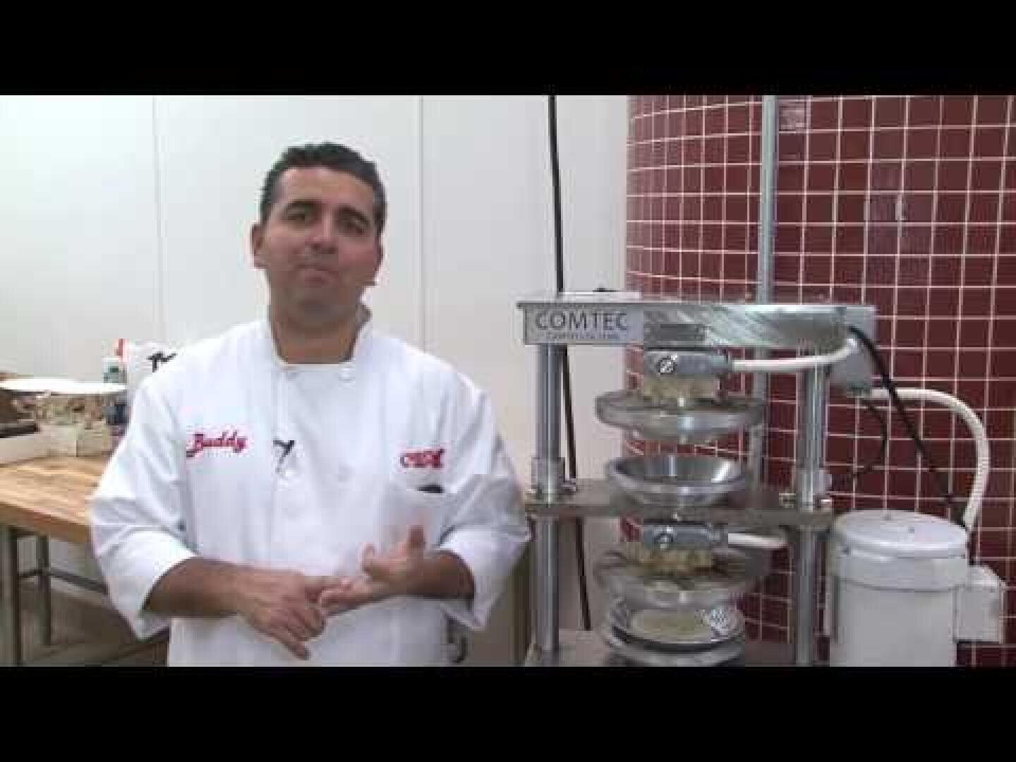 A chef in a white jacket stands next to an industrial pastry press machine in a bakery kitchen with tiled and white walls.