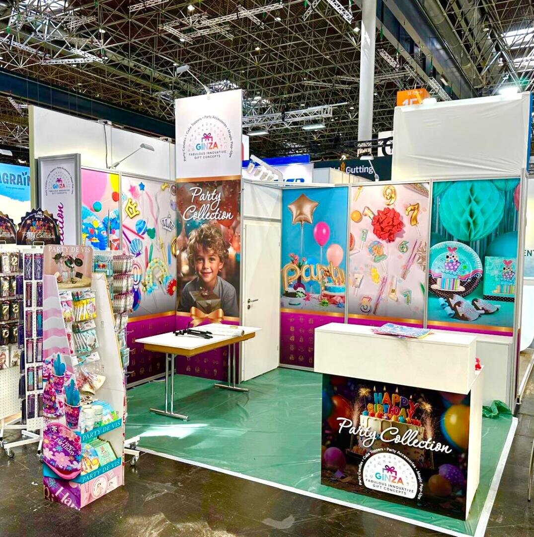 Ein farbenfroher Messestand präsentiert Partyzubehör wie Luftballons, Banner und Dekorationen. Bunte Poster, ein lächelndes Kind und geordnete Produkte sorgen für festliche Atmosphäre. Auf einem Tisch und in Regalen finden sich weitere Partyartikel.