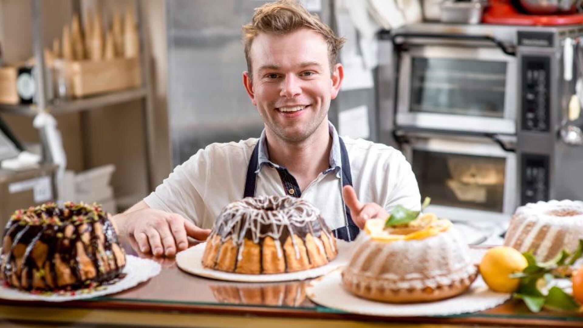 A smiling baker stands behind a counter with four decorated cakes; ovens and kitchen appliances can be seen in the background.