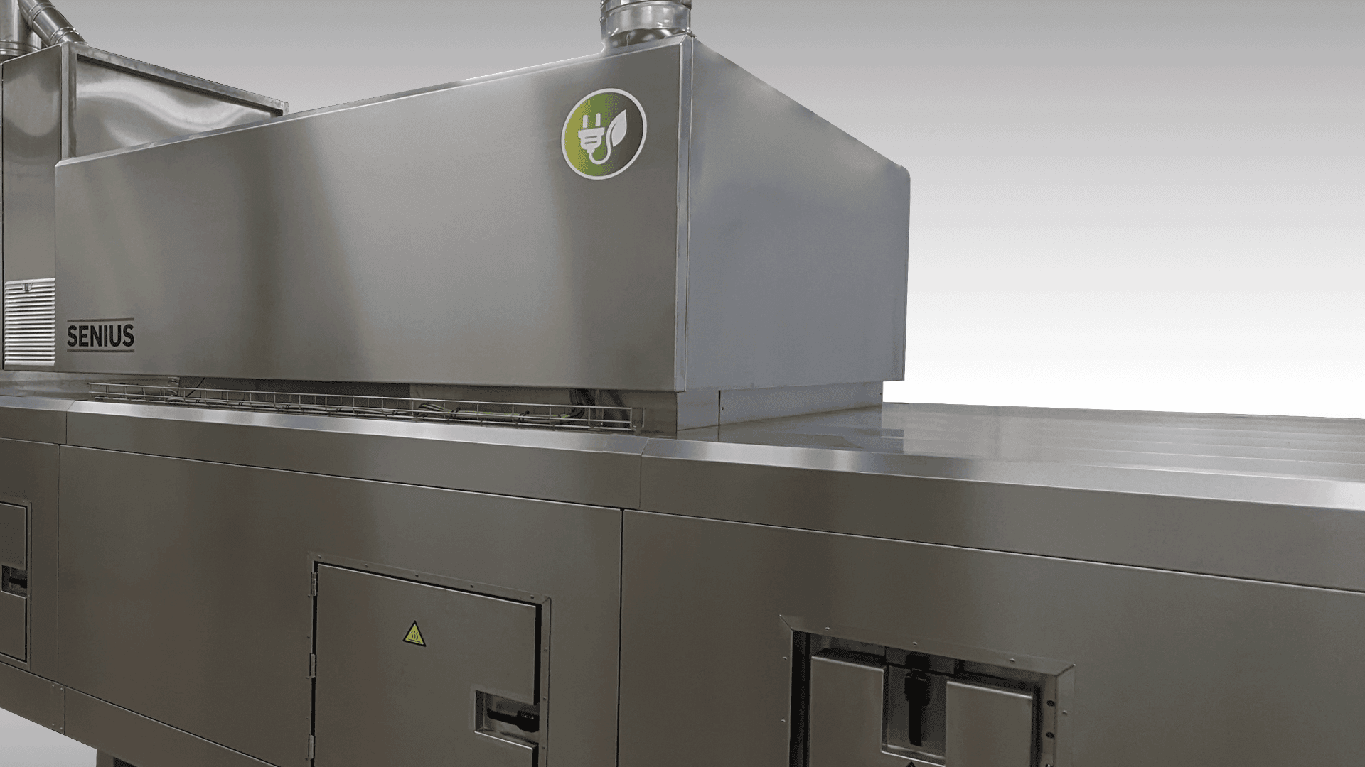 Eine große industrielle Lebensmittelverarbeitungsmaschine aus rostfreiem Stahl mit einem grünen, umweltfreundlichen Aufkleber auf der oberen Einheit, mit der Aufschrift Senius, vor einem schlichten, hellen Hintergrund.
