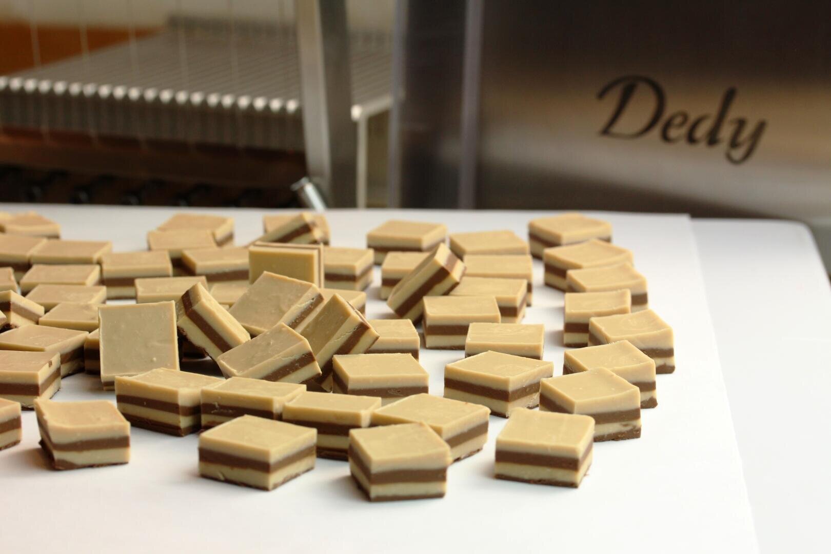 Ein Stapel rechteckiger, geschichteter Pralinen ruht auf weißem Papier. Jedes Stück hat abwechselnd helle und dunkle Schichten. Im Hintergrund ist ein Teil einer Maschine zu sehen, in die das Wort Dedy eingraviert ist.