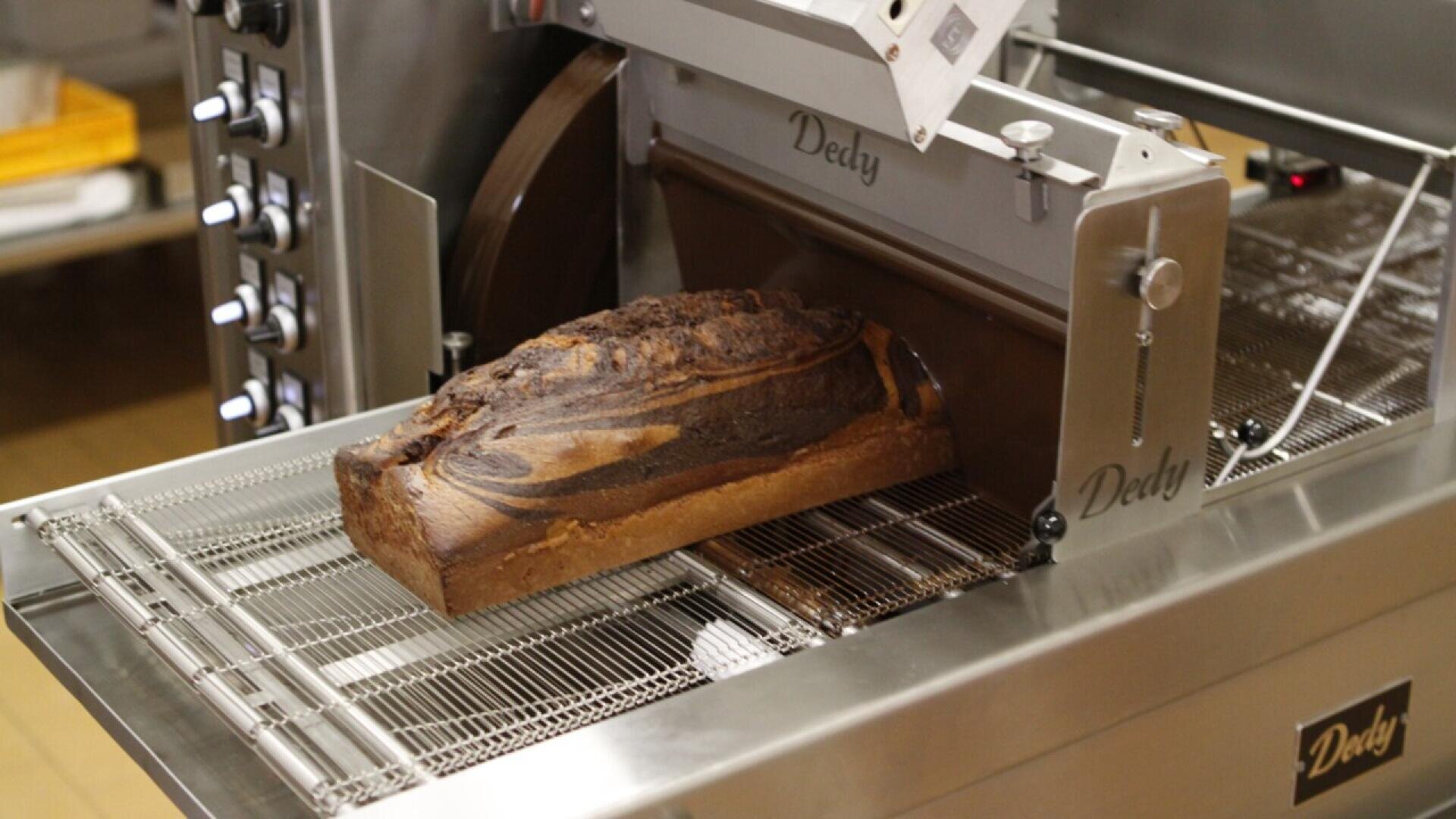 A loaf of marble bread sits on the conveyor of an industrial bread slicer machine in a bakery setting.