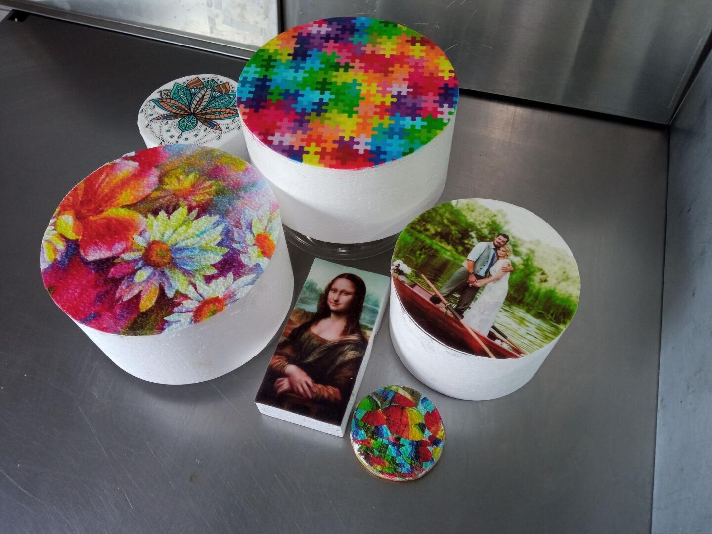 Fünf runde und ein rechteckiger Kuchen stehen auf einer Metalloberfläche, jeder ist mit aufgedruckten essbaren Bildern verziert: Blumen, ein buntes Puzzle, ein Mandala, ein Hochzeitsfoto, die Mona Lisa und ein geometrisches Muster.