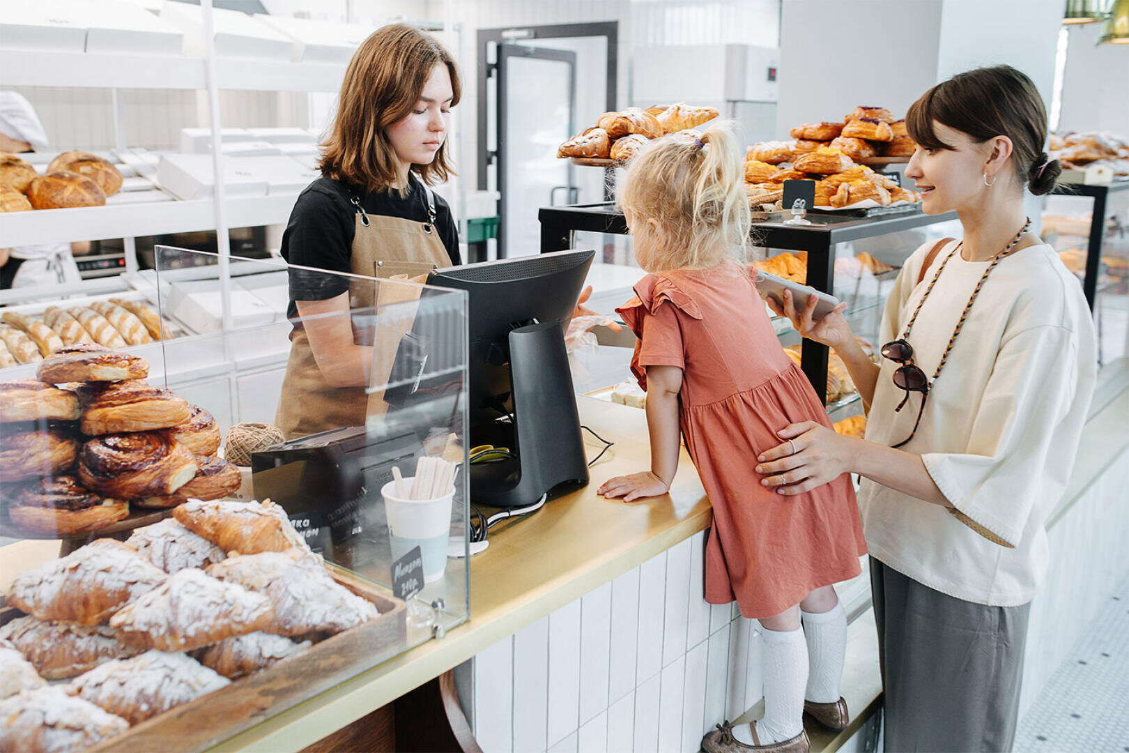 A woman and a young girl stand at a bakery counter, choosing pastries. The girl leans on the counter while an employee in an apron stands behind the register. Display cases are filled with various breads and pastries.