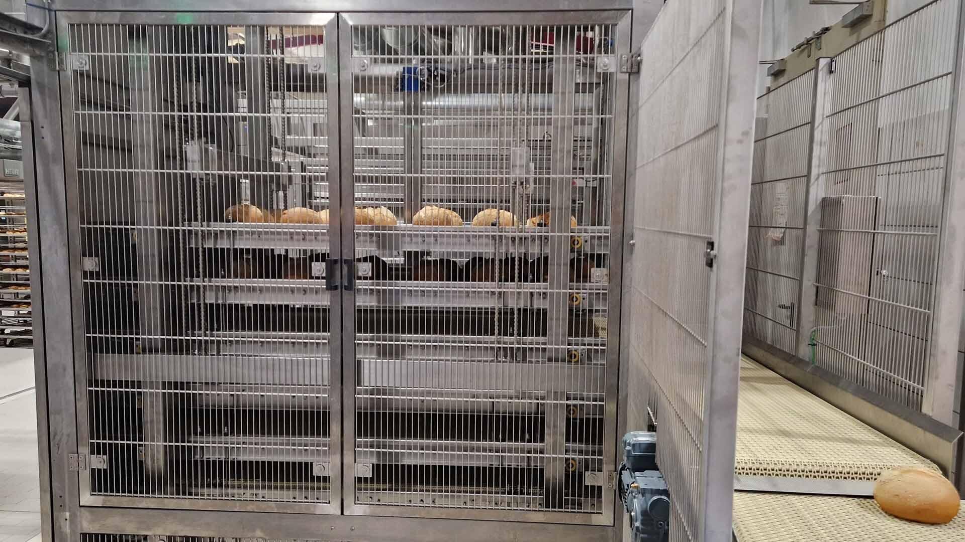 Mehrere Brotlaibe in einem großen industriellen Metallofen mit Gittertüren; ein Laib liegt auf einem Förderband außerhalb des Ofens in einer gewerblichen Backstube.