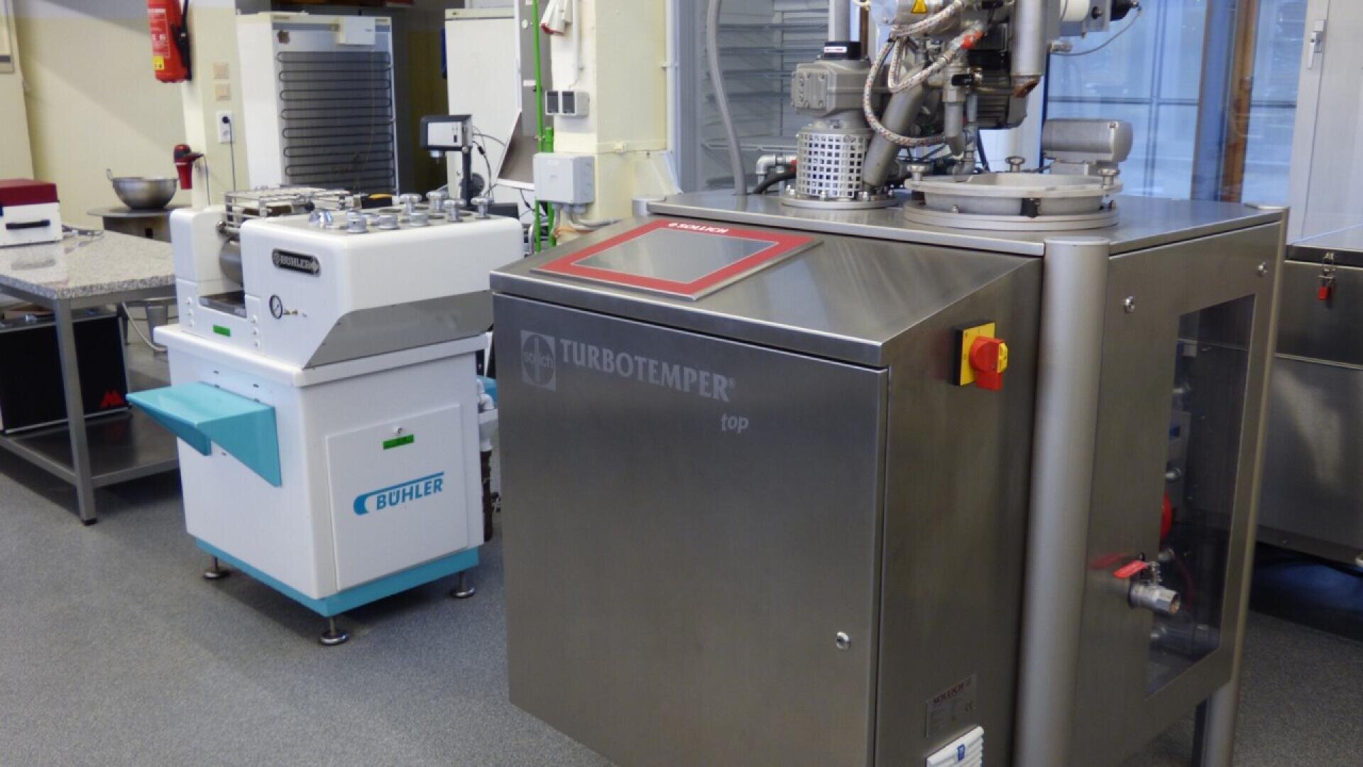 A stainless steel TURBOTEMPER machine and a white BUHLER machine are placed in a clean, modern laboratory with gray flooring and various scientific equipment in the background.