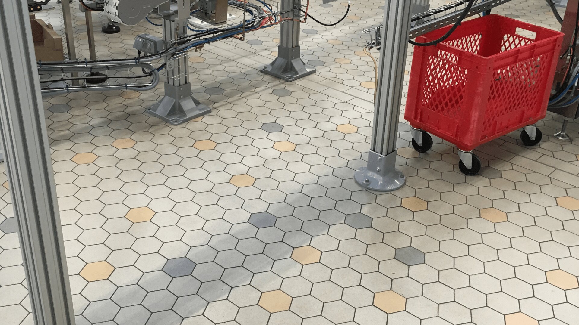 Ein roter Kunststoffkorb auf Rädern steht auf einem sechseckigen gefliesten Boden, umgeben von Metallpfosten und Maschinen mit freiliegenden Kabeln.
