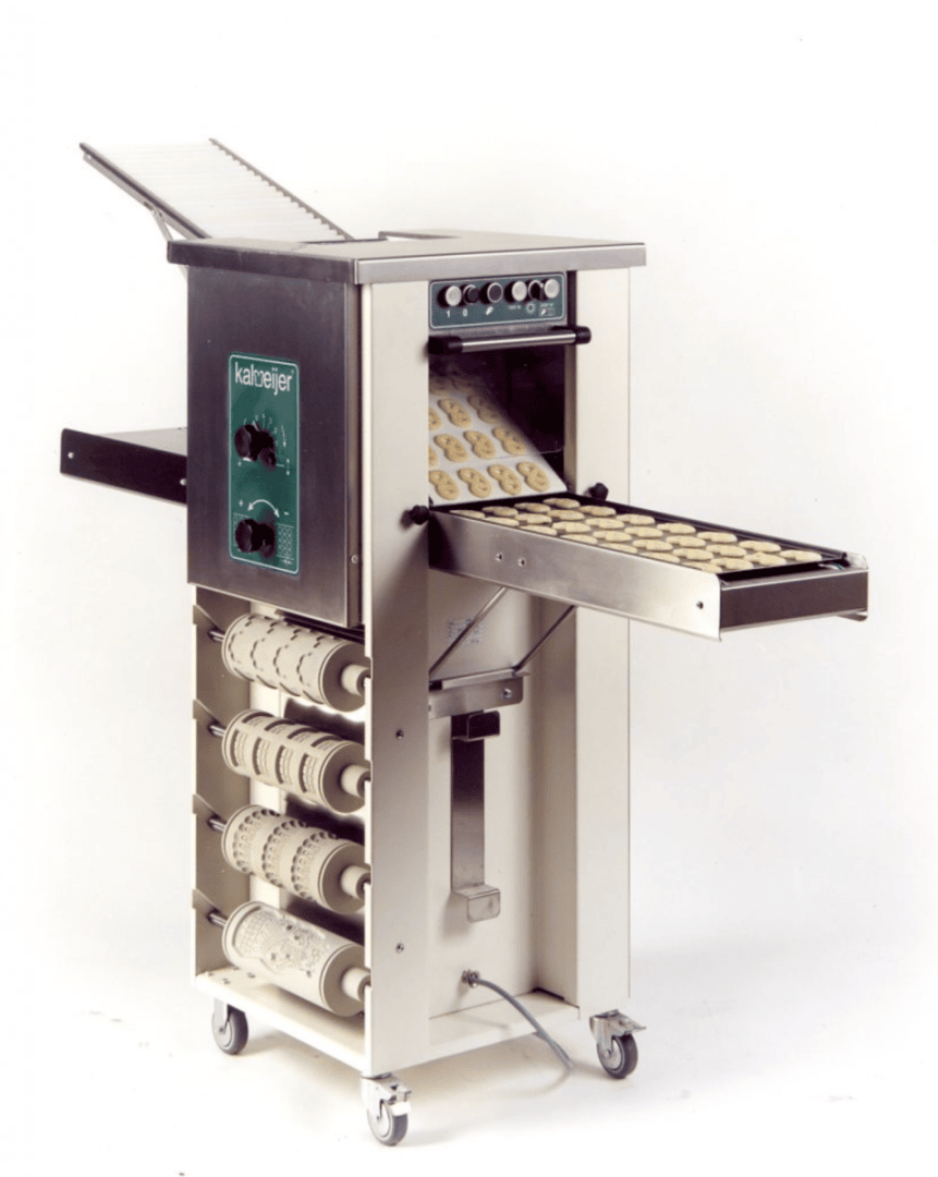 Eine kommerzielle Plätzchenbackmaschine, aus der Teigschalen herauskommen, mit verschiedenen gemusterten Walzen, die an der Seite aufbewahrt werden, und mit Steuerschaltern an der Vorderseite. Die Maschine steht auf Rädern, damit sie mobil ist.