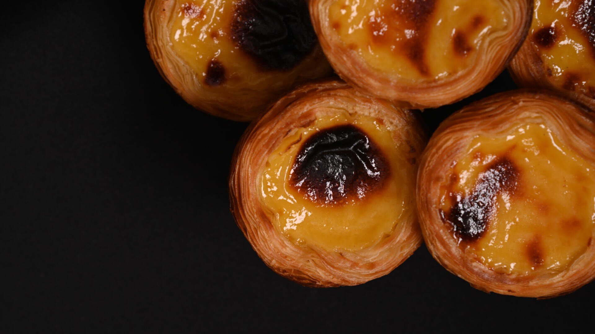 Close-up of several Portuguese custard tarts (pastéis de nata) with flaky, golden-brown crusts and caramelized tops, arranged on a black background.