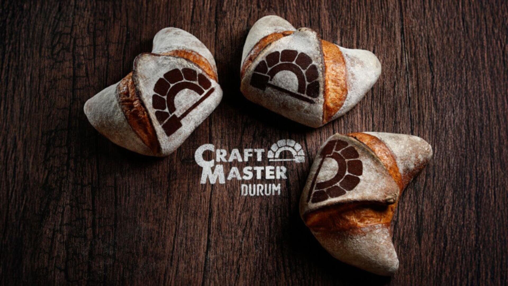 Drei rustikale Brötchen mit einem dunklen Ofenlogo darauf liegen auf einer Holzfläche. Unter dem Brot sind der Schriftzug „Craft Master Durum“ und das gleiche Ofenlogo auf dem Holz angebracht.
