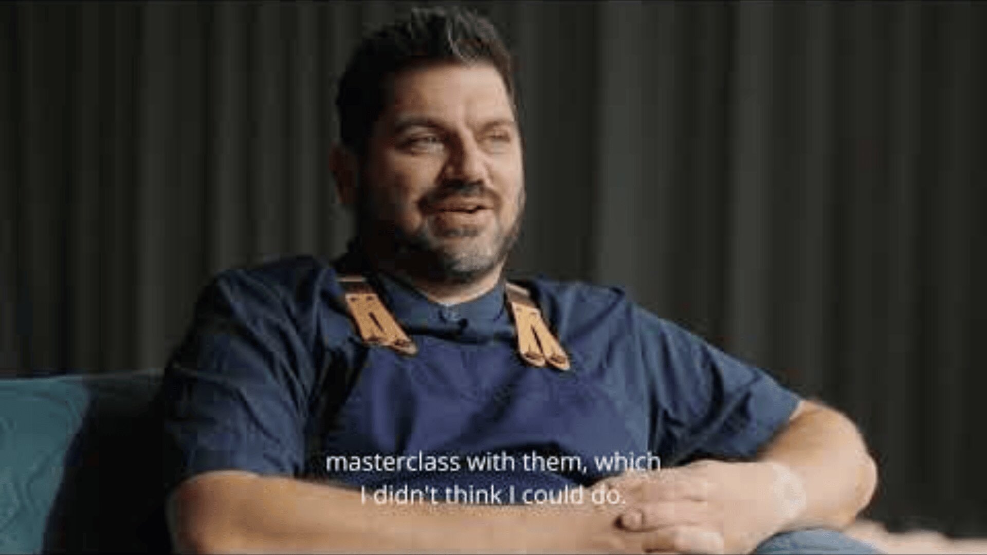 A man with dark hair and a beard, wearing a blue shirt and an apron, sits on a chair and smiles. The background is dark and blurred. The subtitles at the bottom read: Master class with them, which I wouldnt have thought possible.