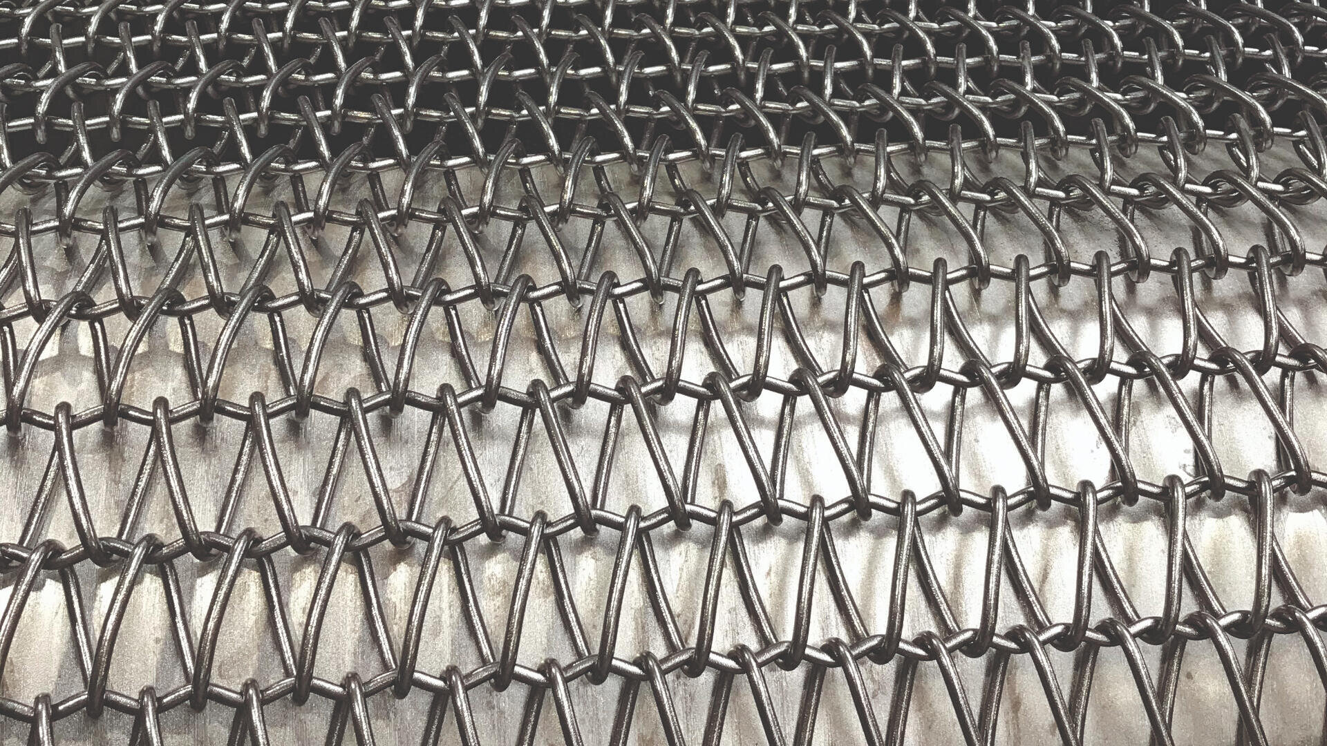 Nahaufnahme von verflochtenen Metalldrähten, die auf einer zylindrischen Metalloberfläche ein kettenartiges Netzmuster bilden. Das Netz bildet eine sich wiederholende Rautenform.