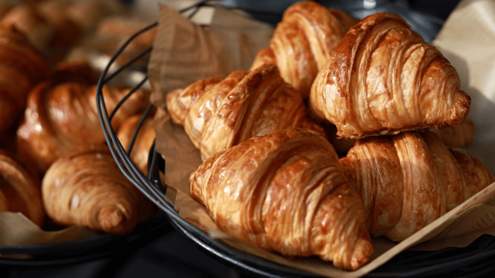 A basket lined with brown parchment paper is filled with golden, flaky croissants. More croissants are visible in a basket in the background. The pastries appear fresh and richly browned.