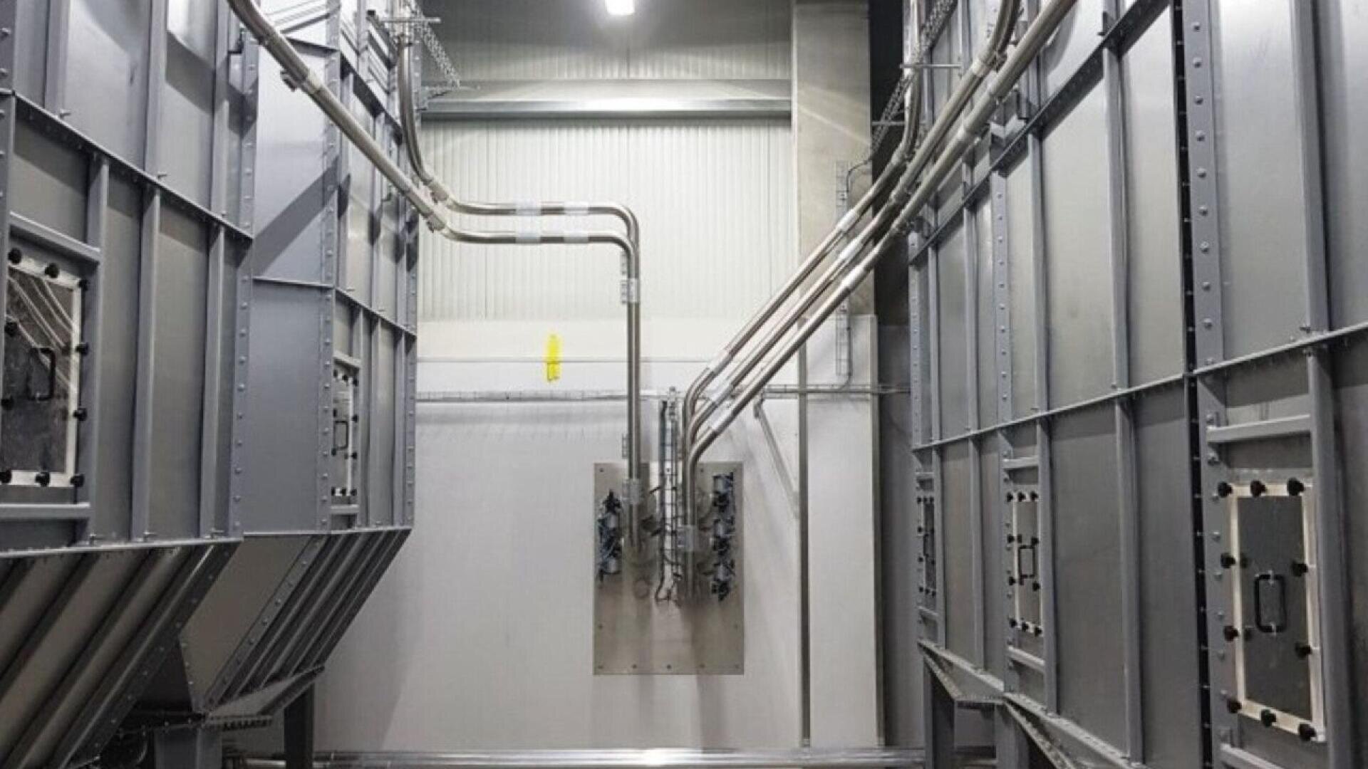 A view of an industrial facility interior showing large metal structures, vertical metal panels, and several pipes and cables connected to a central wall-mounted electrical panel. The area is well-lit with overhead lighting.