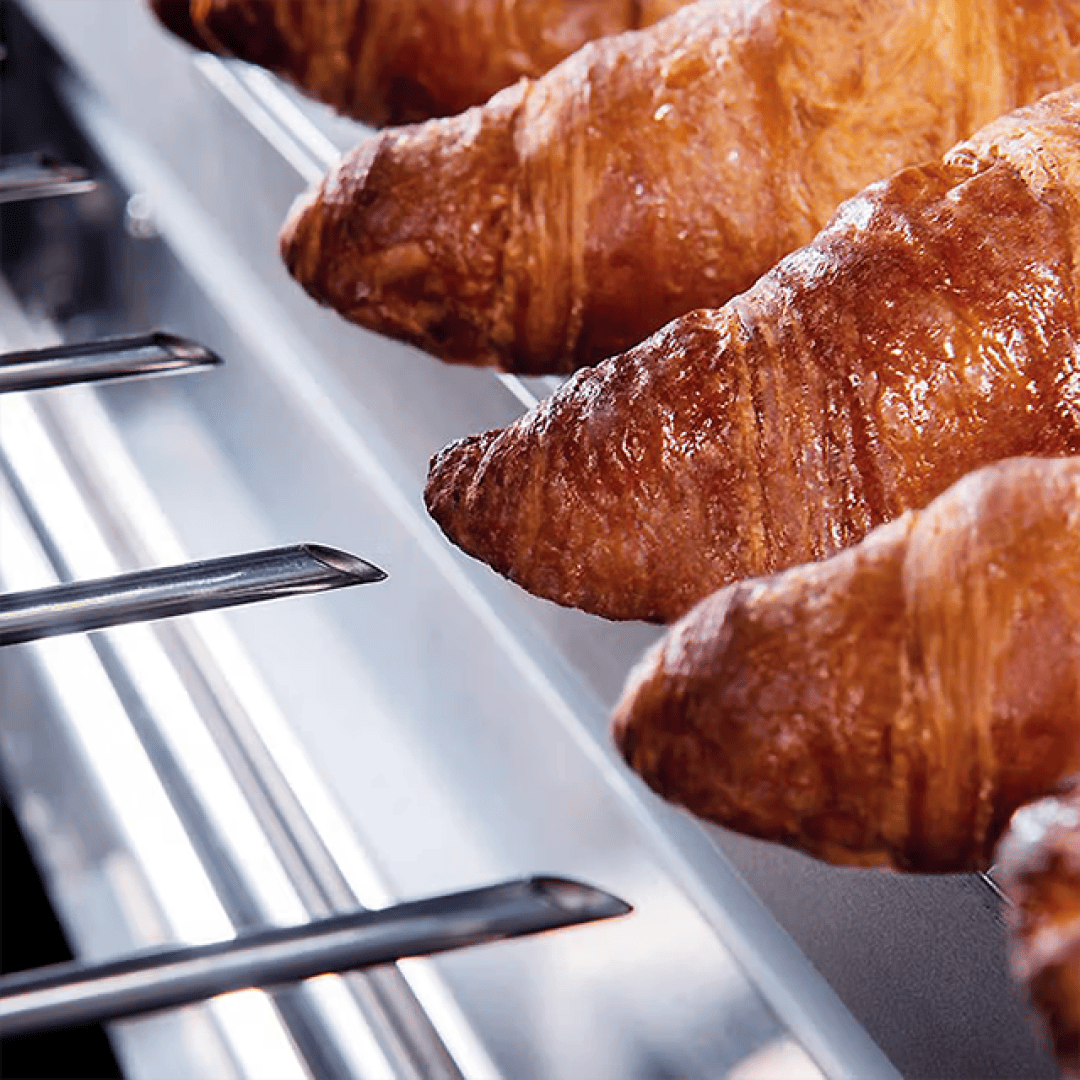 Several golden-brown croissants are lined up on metal rods in a display case or bakery, showing off their flaky texture and shiny, glazed surface.