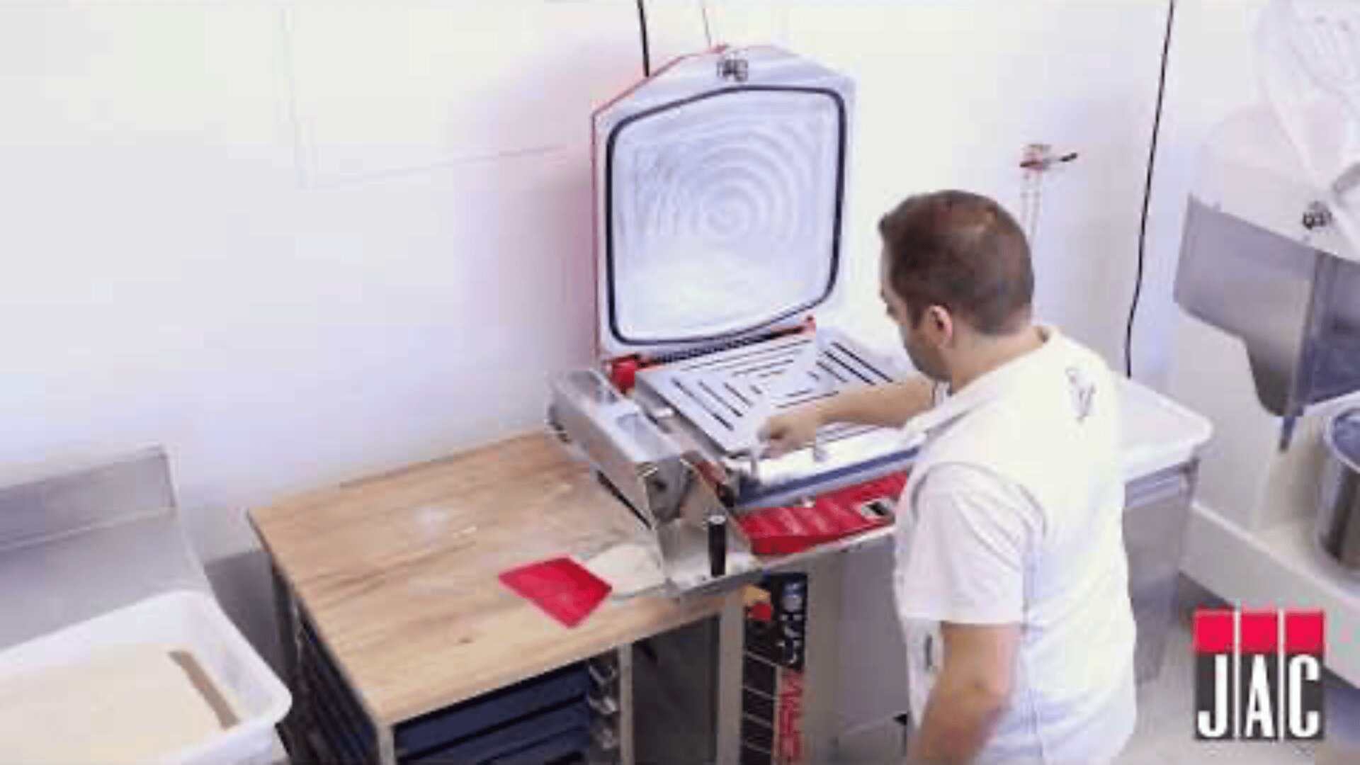 A man in a white shirt operates a commercial vacuum sealing machine in a clean, industrial kitchen setting. The machine lid is open, and the JAIC logo is visible in the bottom right corner.