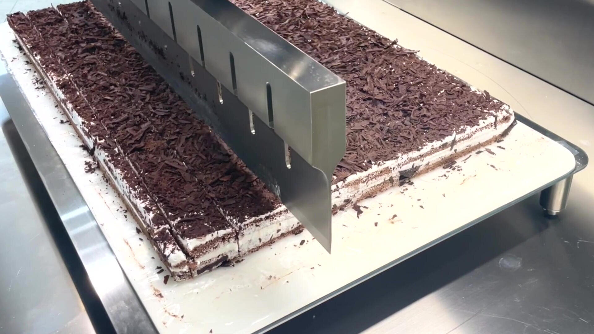 A large rectangular cake with chocolate shavings on top is being sliced into even pieces by an industrial metal cake cutter in a commercial kitchen setting.