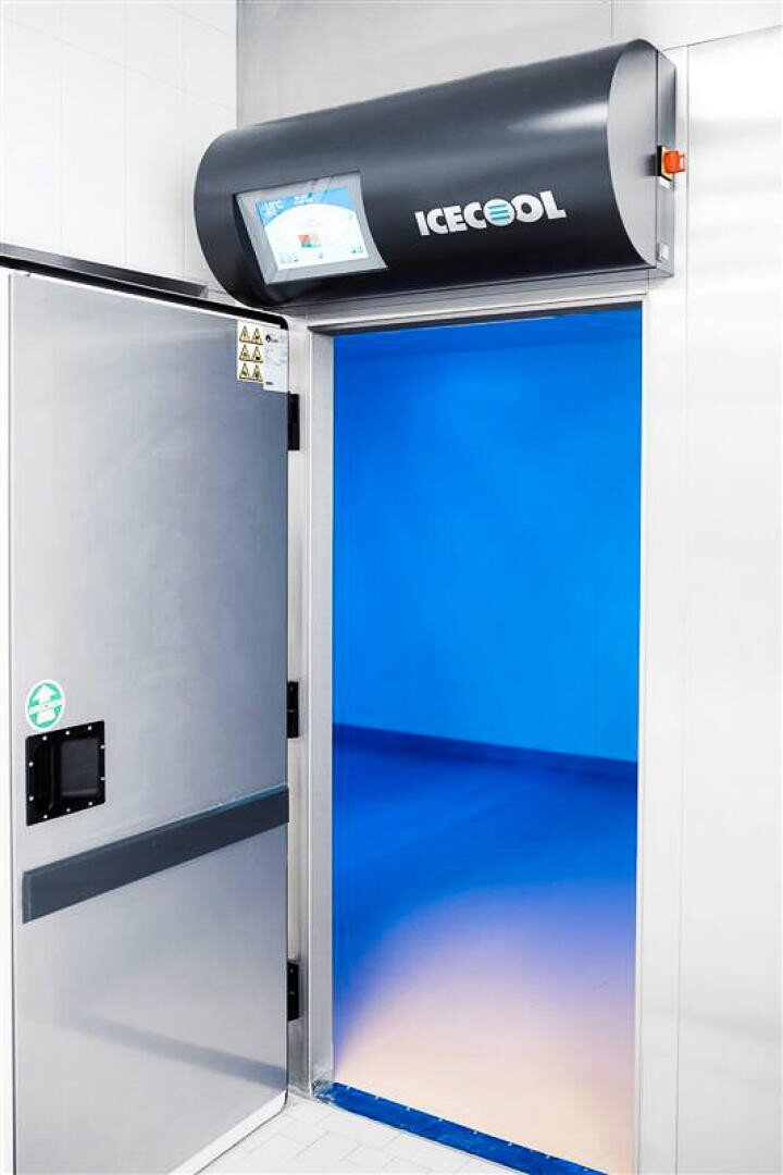 Ein modernes industrielles Kühlhaustor mit einem blau isolierten Eingang, darüber die Aufschrift ICECOOL. Auf der linken Seite befindet sich eine teilweise geöffnete Metalltür mit Sicherheitswarnschildern.