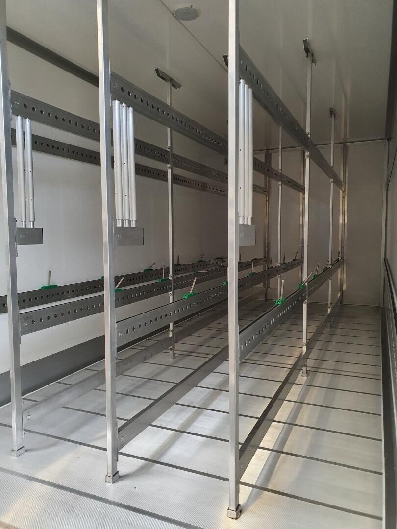 Leere Metallregale mit vertikalen und horizontalen Stangen sind in einem sauberen, gut beleuchteten, weißen Raum aufgestellt. Die Regale scheinen zur Aufbewahrung oder zum Aufhängen bereit zu sein; an den Stangen sind grüne Haken angebracht.