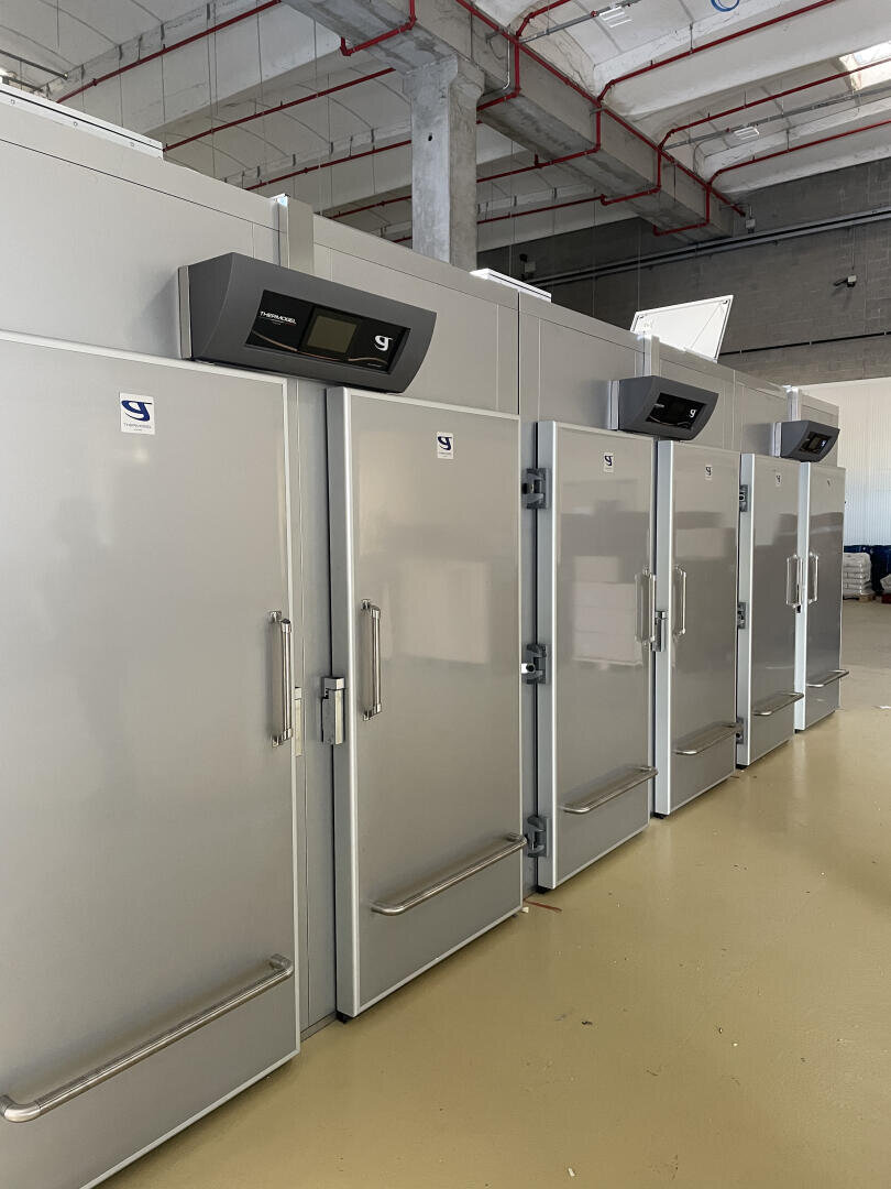 A row of large industrial refrigerators or freezers with digital displays stands in a spacious room with high ceilings and exposed pipes. The units are grey with vertical handles and are lined up side by side.