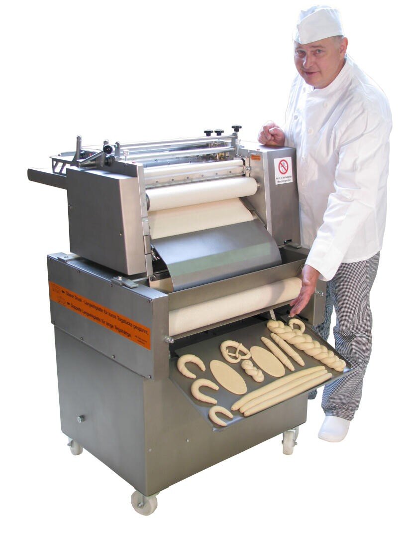 Ein Bäcker in weißer Uniform steht neben einer Teigformmaschine und zeigt verschiedene Formen von rohen Teiglingen, die auf einem Tablett angeordnet sind, das aus der Maschine herausragt.