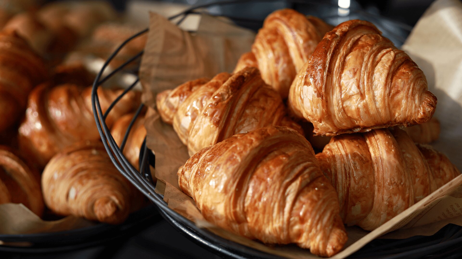 Eine Nahaufnahme von goldbraunen, blättrigen Croissants, die in einem mit braunem Pergamentpapier ausgelegten Korb gestapelt sind. Im Hintergrund sind weitere Croissants zu sehen.