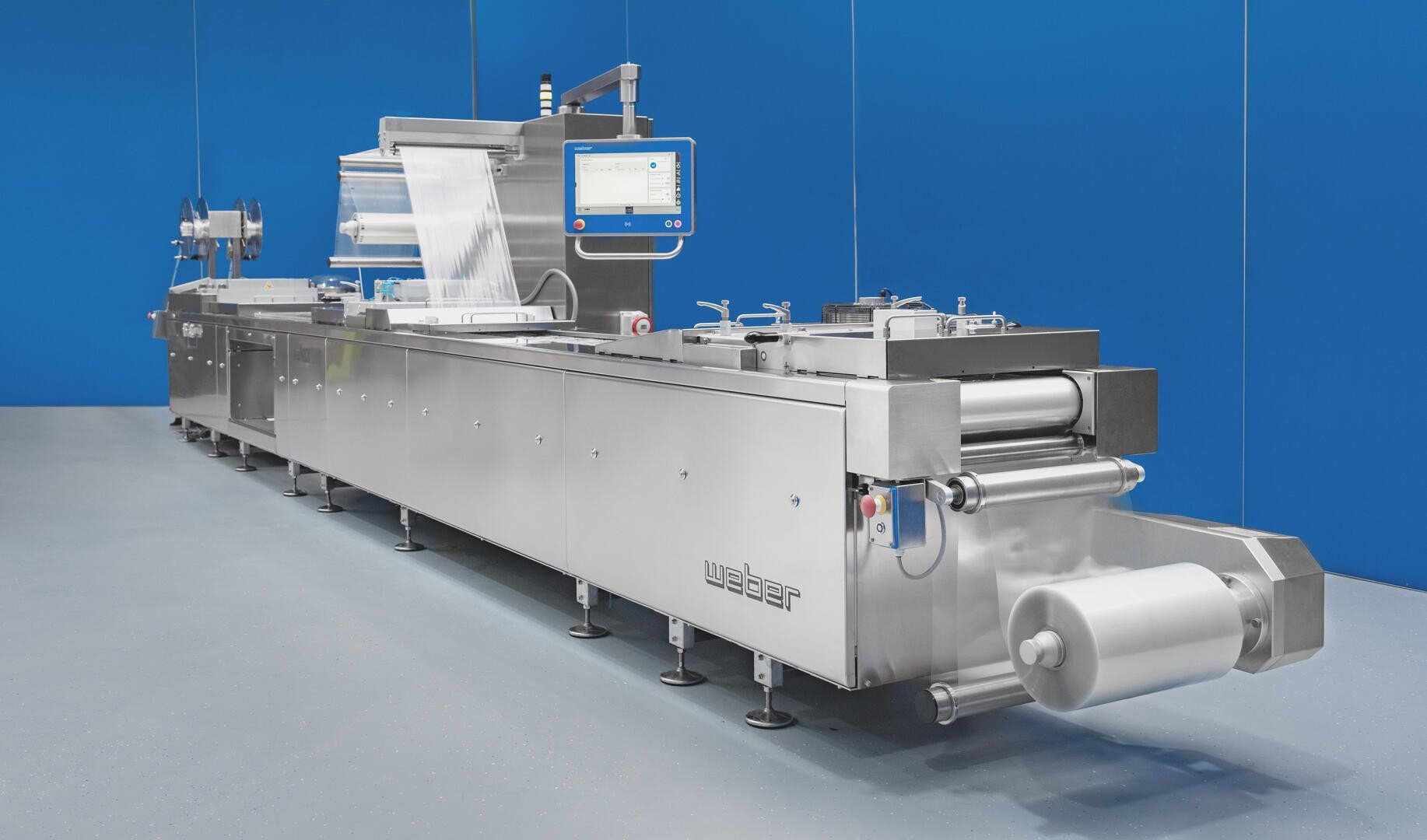 Eine große industrielle Lebensmittelverpackungsmaschine mit einem Gehäuse aus Edelstahl und einem Touchscreen-Display vor einer blauen Wand in einer sauberen, modernen Einrichtung.