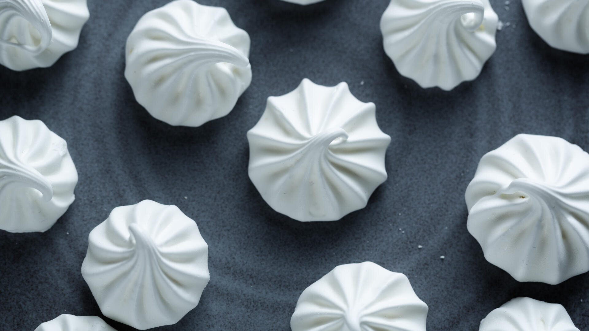 Kleine, weiße Baiser-Kekse mit einer gewirbelten, geriffelten Textur sind auf einer dunklen, strukturierten Oberfläche angeordnet.