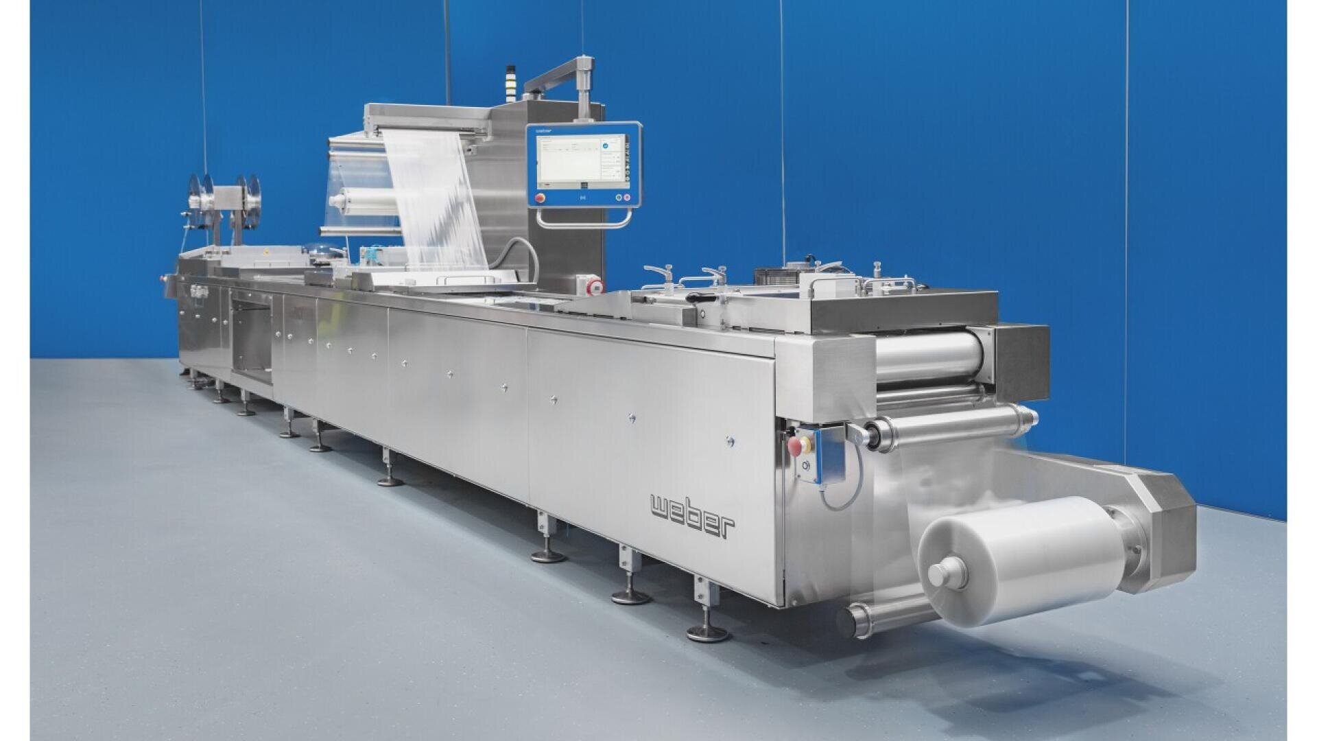 A large industrial food packaging machine made of stainless steel with a touch screen display, clear plastic film, and conveyor belts, set against a blue wall in a clean factory environment.