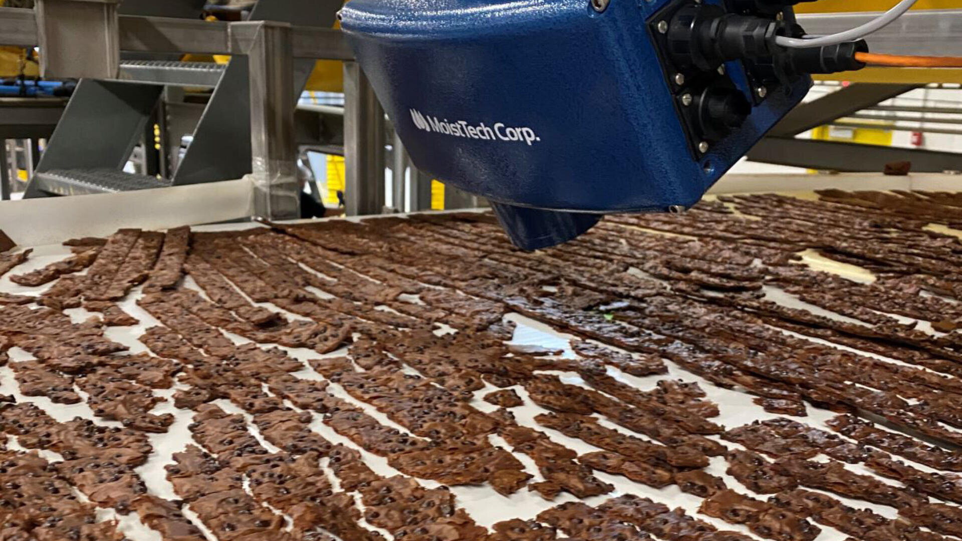 A close-up view of a robotic machine inspecting rows of cooked bacon strips on a conveyor belt in a food processing facility.
