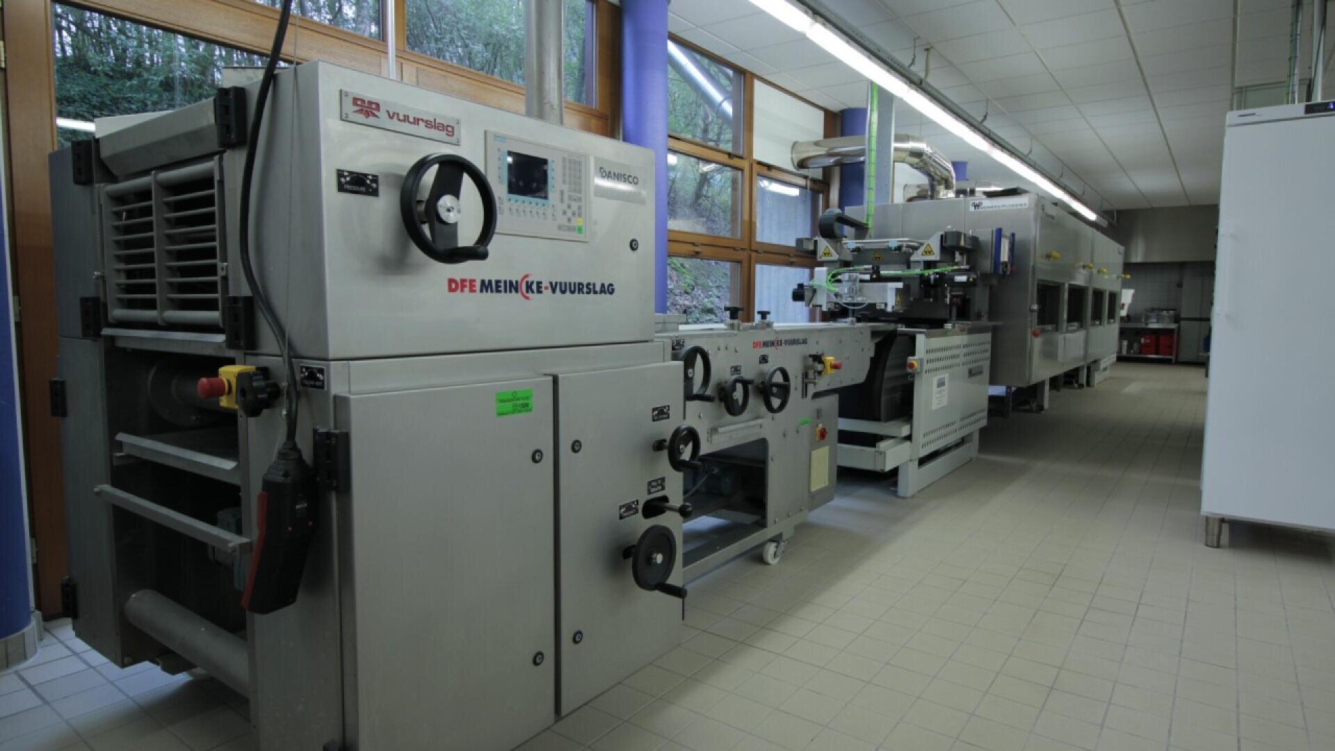 Industrial printing machines with control panels and rollers are lined up in a clean, well-lit facility with large windows and tiled floors.