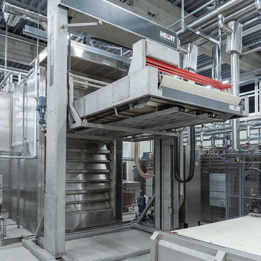Großer industrieller Bäckereiofen mit mehreren Regalen, beschriftet mit HEUFT, in einer Fabrikumgebung mit Metallrohren und Maschinen im Hintergrund. Der Ofen scheint automatisiert zu sein und ist aus rostfreiem Stahl gefertigt.