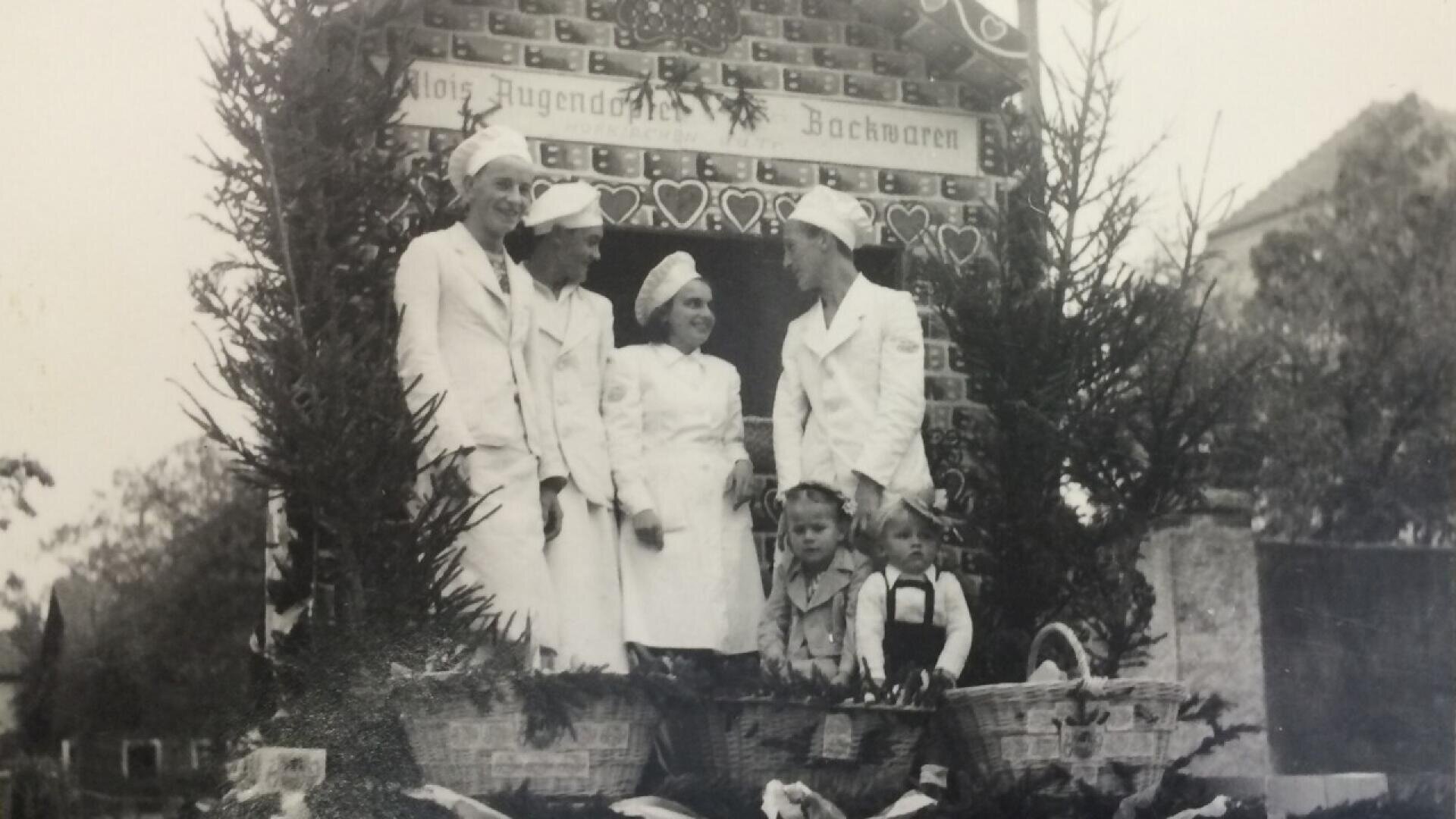 A black and white photo of five people dressed in white uniforms, likely bakers, standing on a decorated float with two children sitting in front. The float is adorned with greenery and a sign reading Alois Jugends Backwaren.