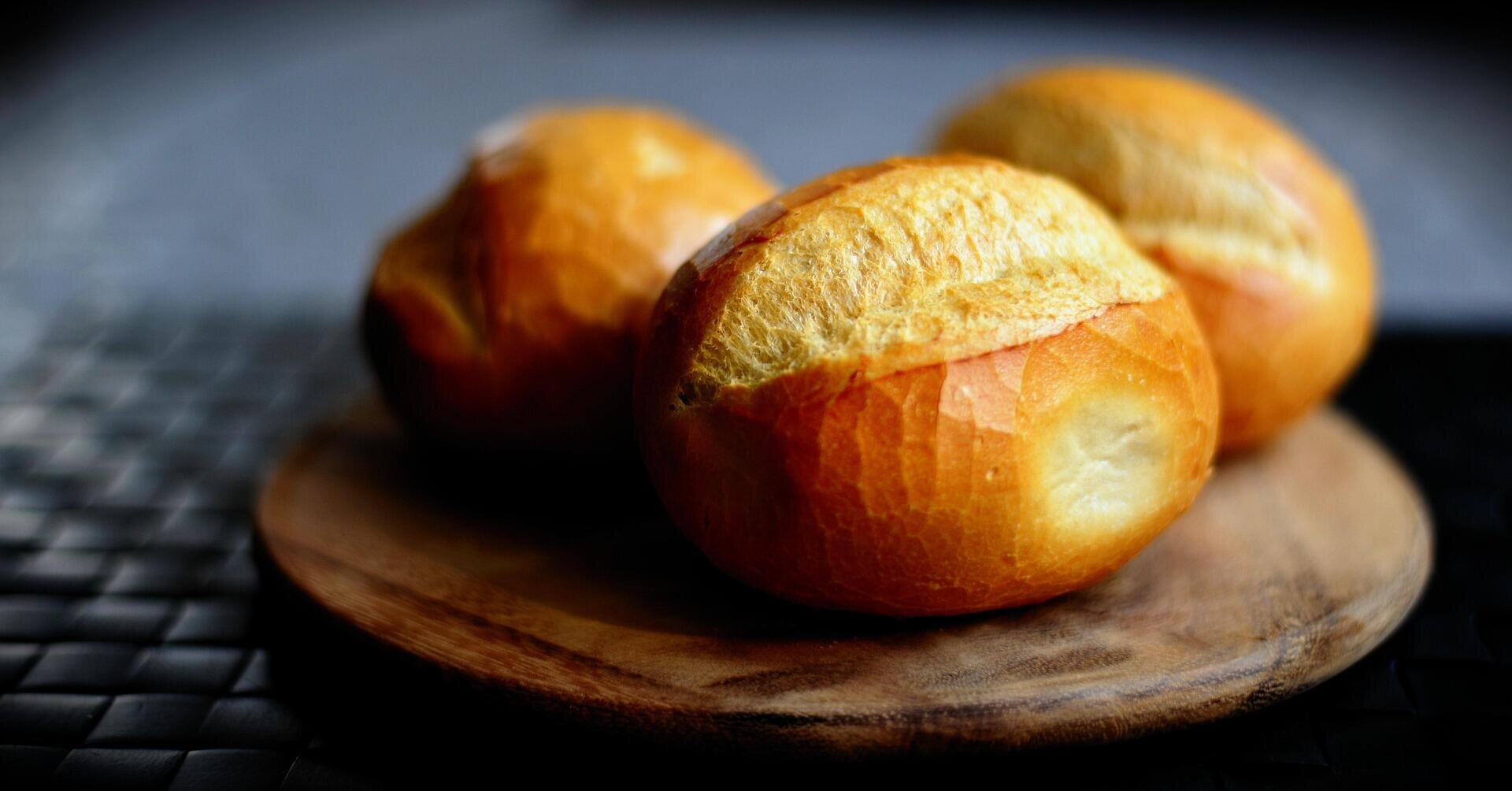 Three round, golden brown bread rolls sit on a small wooden plate. The rolls have crisp, shiny crusts with a split on top and are set against a dark, softly blurred background.