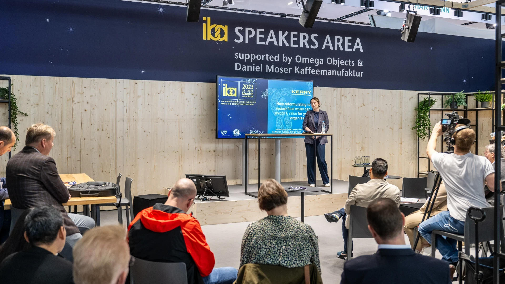 A woman gives a presentation on stage in a Speakers Area at a conference, while an audience listens and photographers take pictures. A large screen beside her displays information about her talk.