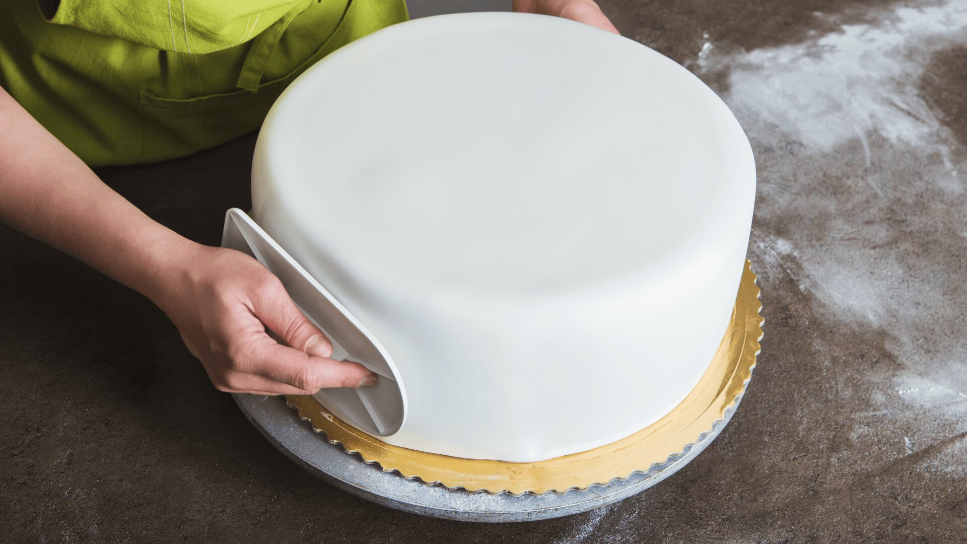 A person smooths white fondant over a round cake on a gold cake board, using a plastic smoother, with flour scattered on the dark countertop.