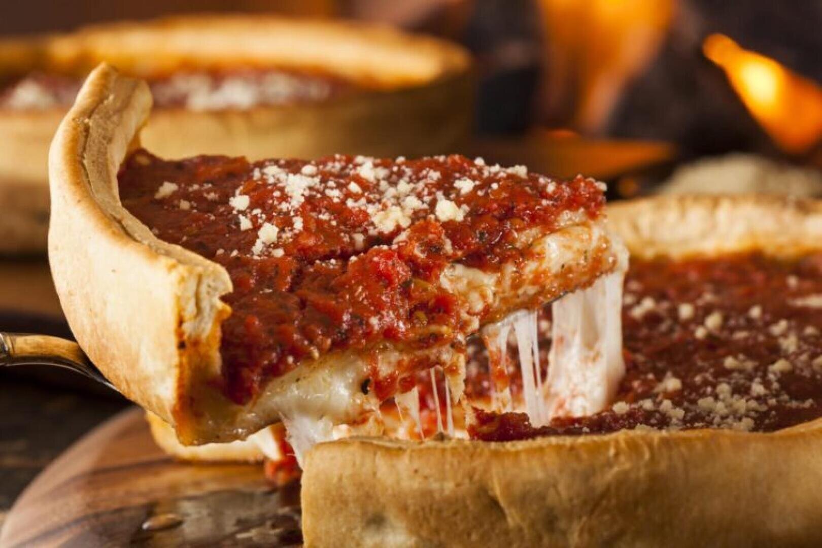 A slice of deep-dish pizza being lifted from the pan, with melted cheese stretching from the crust and chunky tomato sauce on top. The background shows more pizza and a warm, out-of-focus setting.