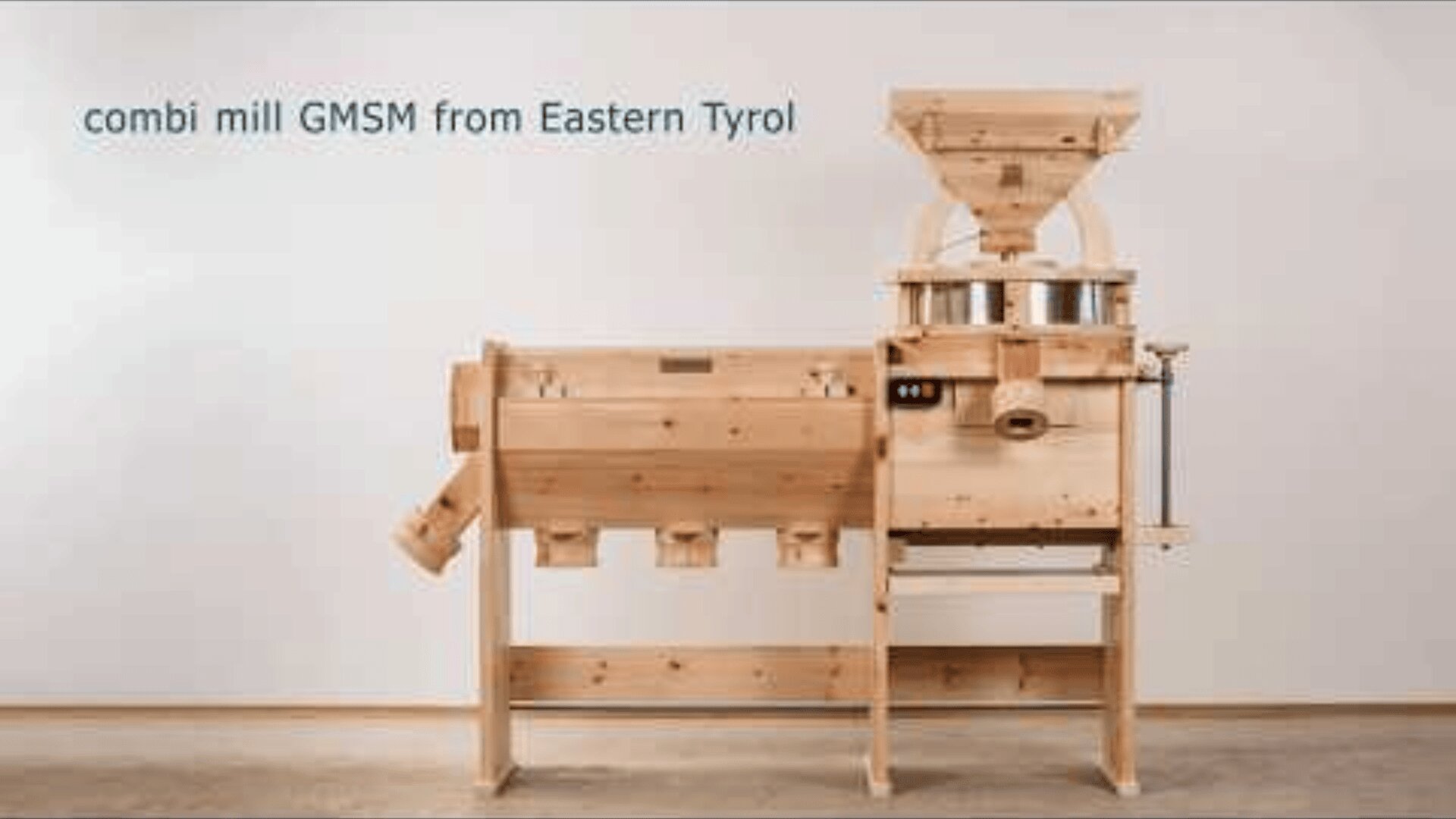 A large wooden combi mill labeled GMSM from Eastern Tyrol stands against a plain wall, showcasing its hopper, grinding stones, and collection compartments.