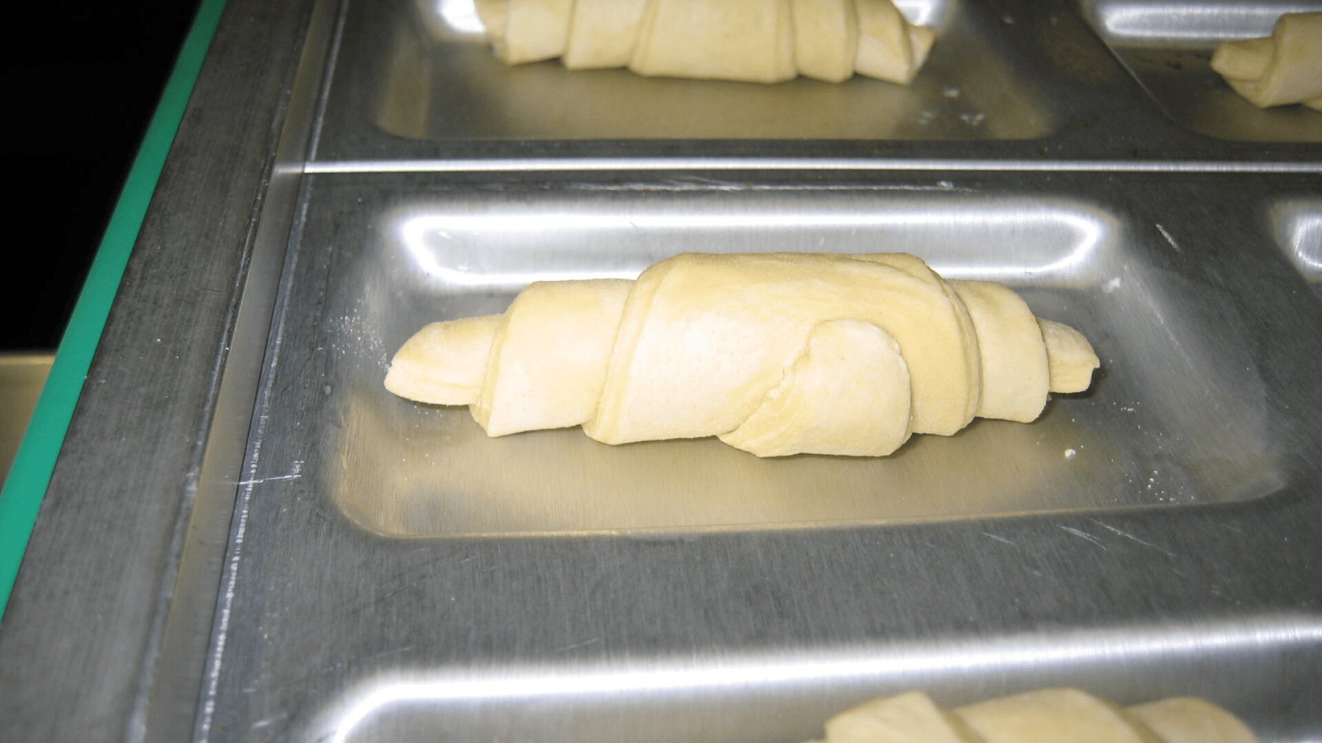 A close-up of an unbaked croissant roll on a metal baking tray, with other unbaked croissants visible in the background.