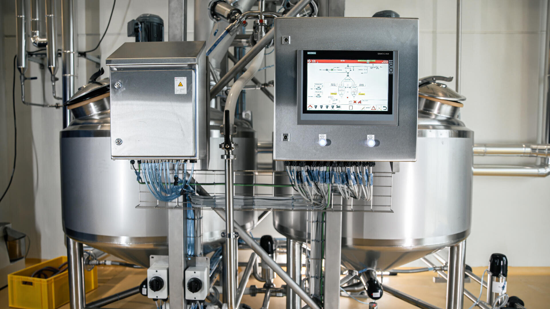 Industrial control panel with a touchscreen display showing a process diagram, positioned in front of stainless steel tanks and various pipes in a modern manufacturing or processing facility.