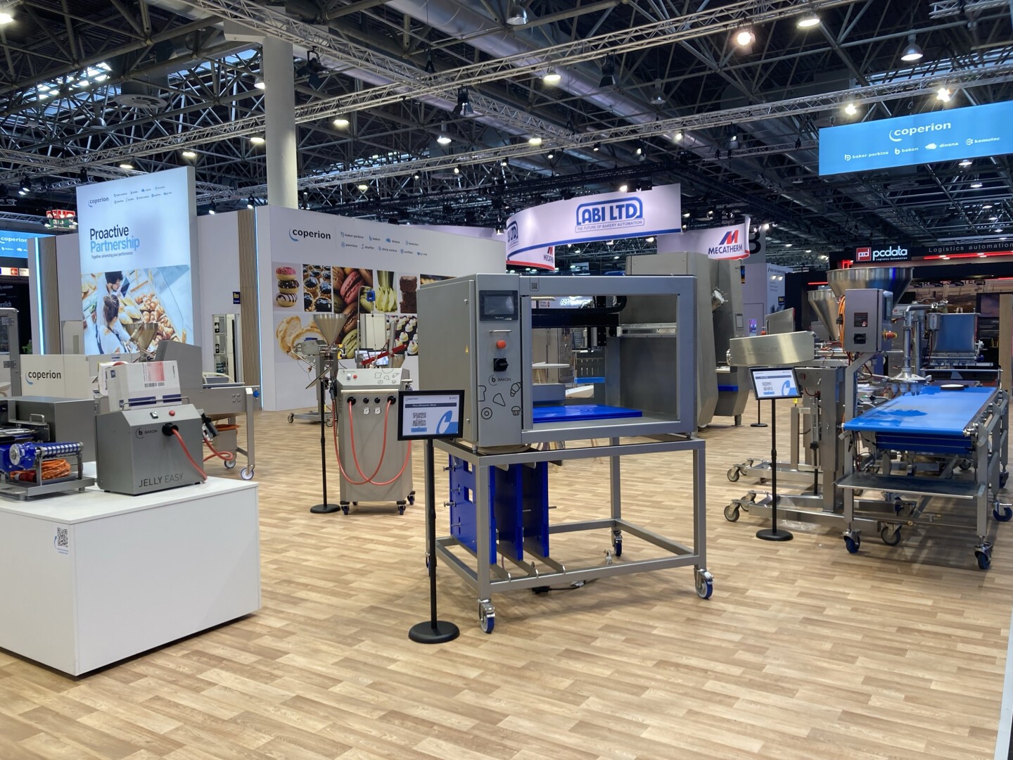 A trade show booth displays various industrial food processing machines on a wooden floor, with bright overhead lighting and company banners, including one for Coperion, in the background.
