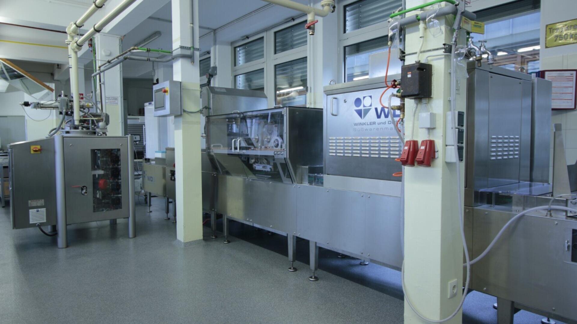 A modern industrial food processing and packaging machine is set up in a clean, well-lit factory environment with metal surfaces and large windows in the background.