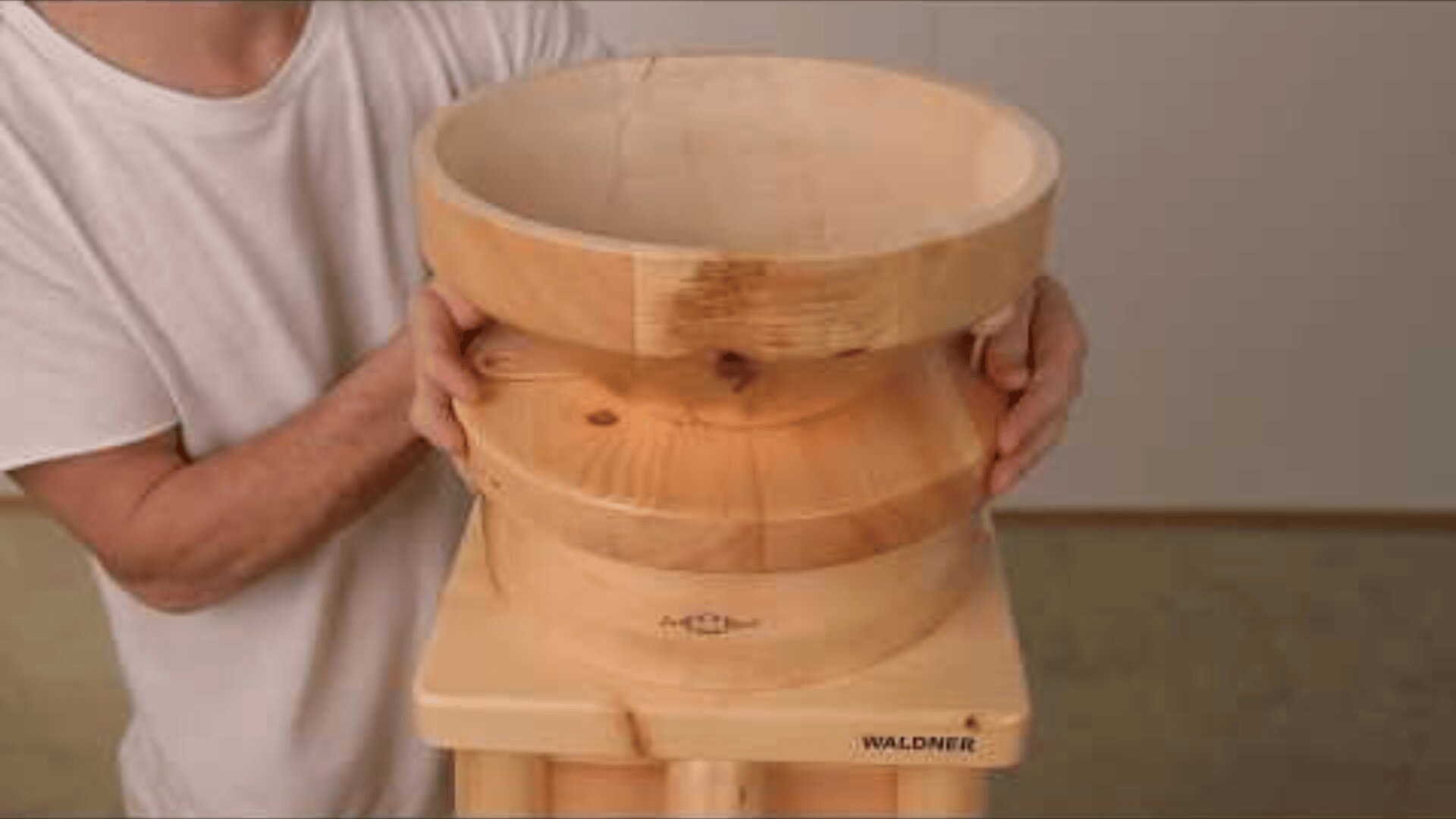 A person in a white shirt holds a large, round wooden bowl attached to a wooden base labeled WALDNER indoors, with a plain background.