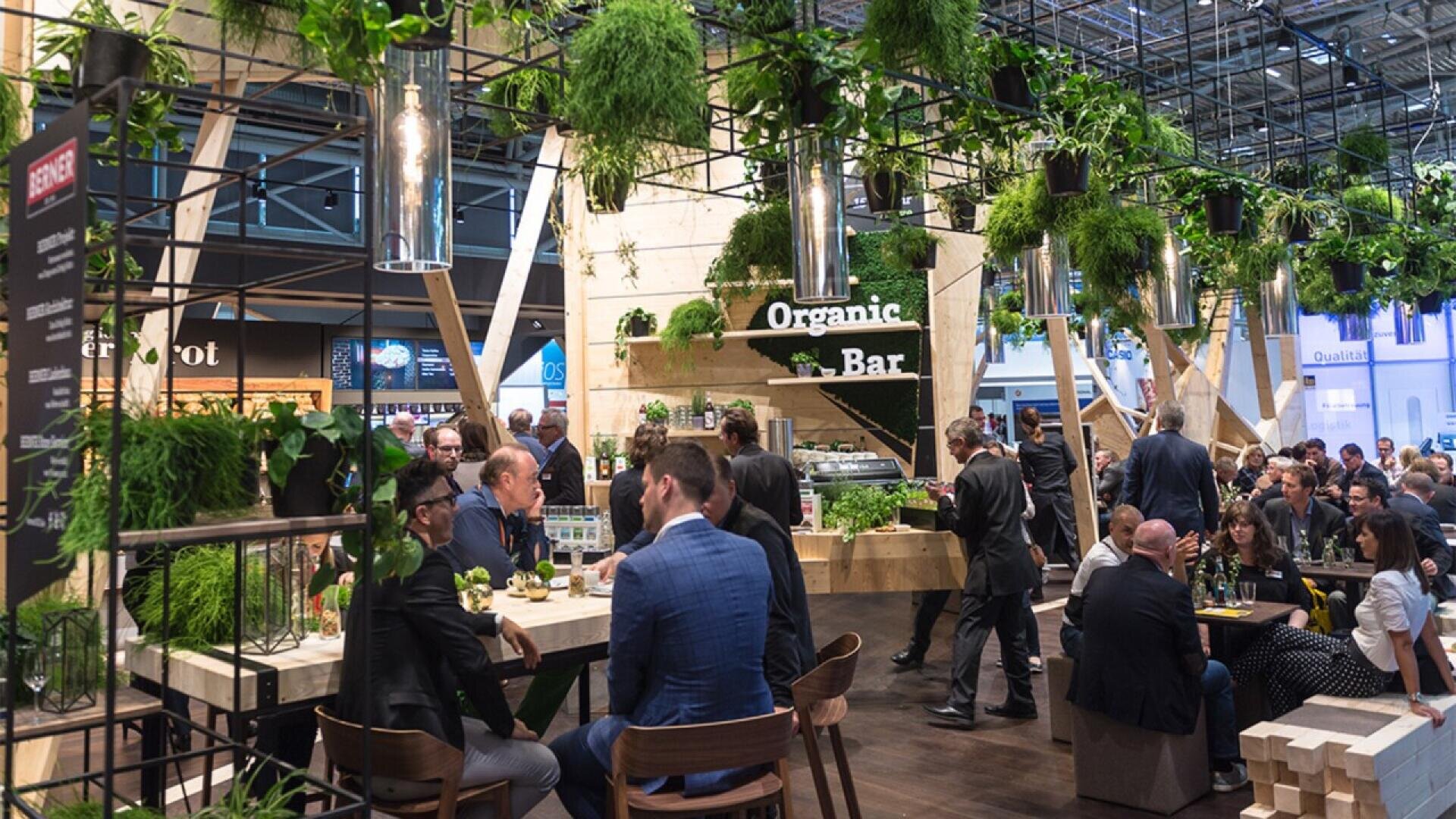 A lively indoor café with many people sitting at tables and socializing. The space is filled with hanging plants and greenery, and a sign reads Organic Bar. The atmosphere is modern and bustling.