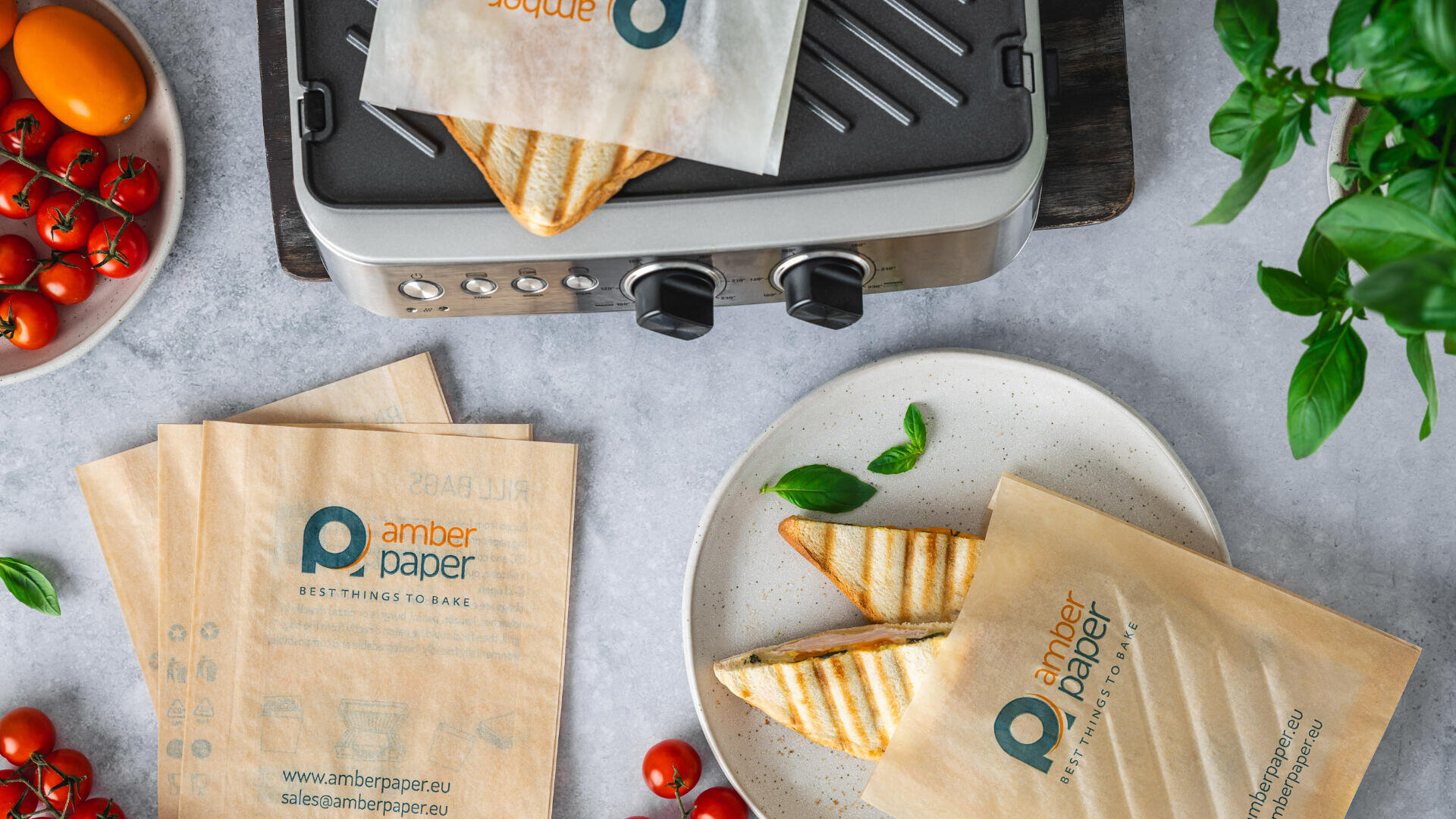 A tabletop grill toasts sandwiches in brown paper sleeves labeled amber paper, with fresh tomatoes, basil, and more paper sleeves on a gray surface. Toasted sandwiches rest on a white plate.