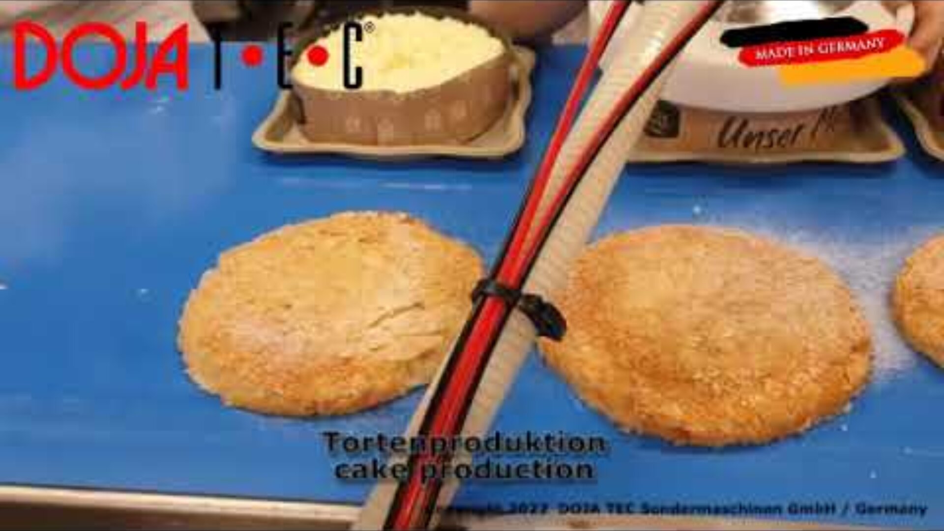Two round cake bases on a blue conveyor belt are being processed in a bakery. A tray of grated cheese or topping is in the background. The DOJA TEC logo and Made in Germany label are visible on the image.