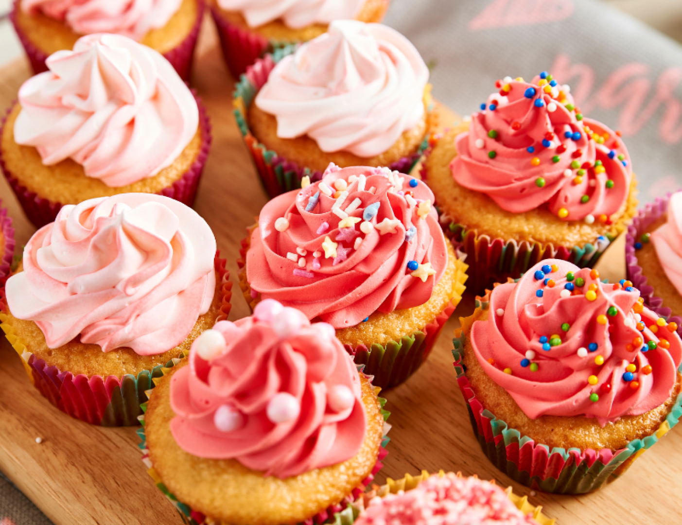 A selection of cupcakes with pink and red icing, some sprinkled with colorful sprinkles, lined up tightly on a wooden surface.