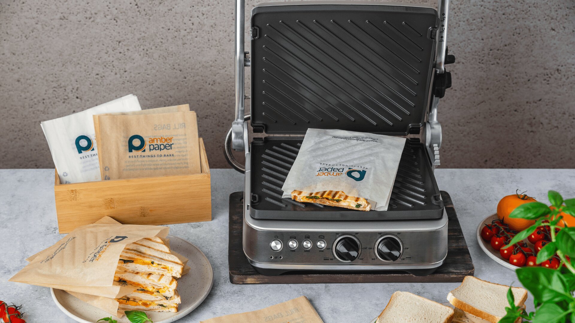 Eine Panini-Presse grillt ein Sandwich in einer Papiertüte mit Markenzeichen. Zusätzliche Papiertüten, aufgeschnittene Sandwiches, Brot, Tomaten und Basilikum sind auf der Arbeitsplatte angeordnet.