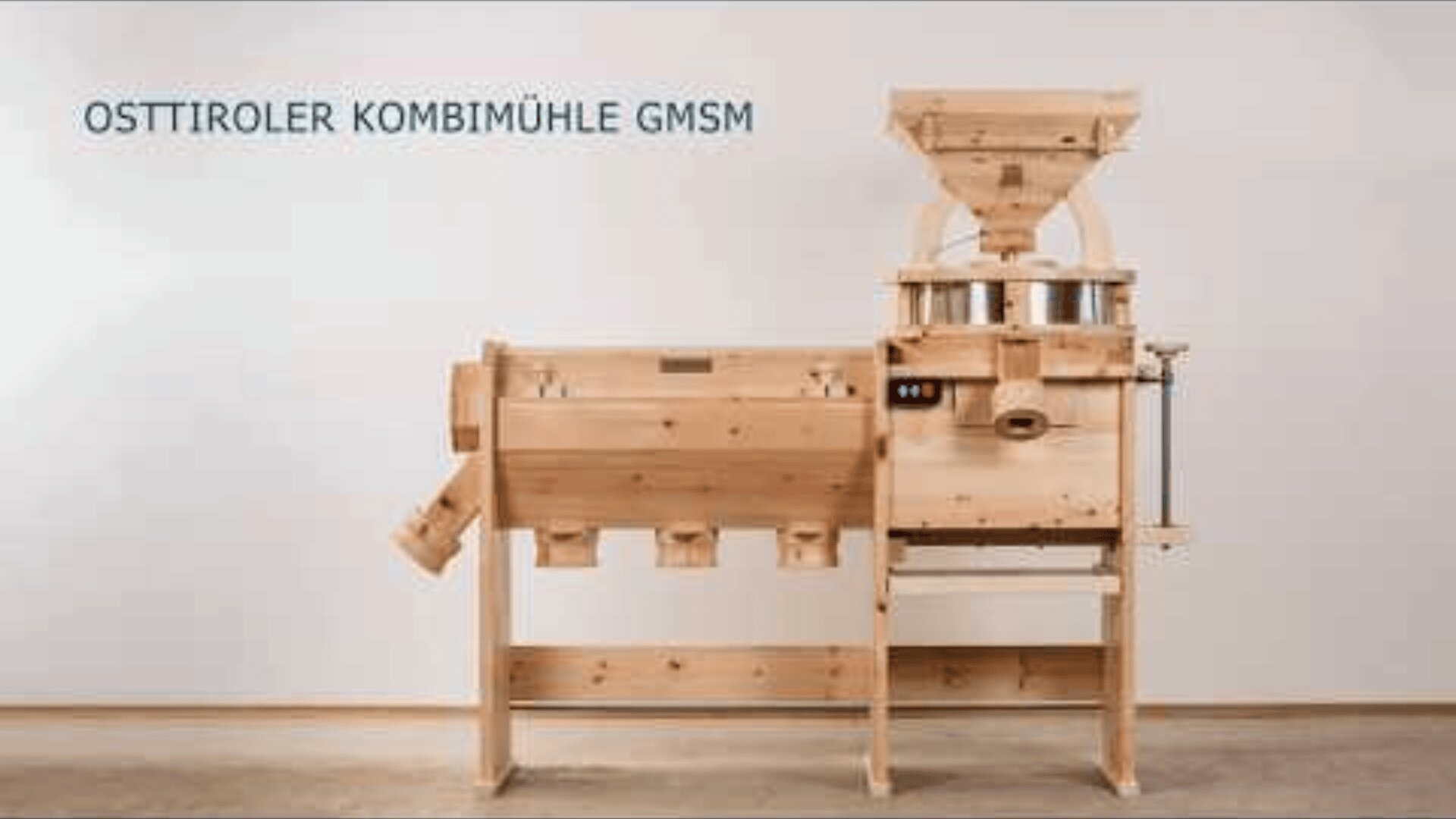 Eine hölzerne Getreidemühle mit der Aufschrift OSTTIROLER KOMBIMÜHLE GMSM steht an einer schlichten Wand und ist mit einem Trichter, einem Mahlwerk und mehreren Auffangbehältern ausgestattet.