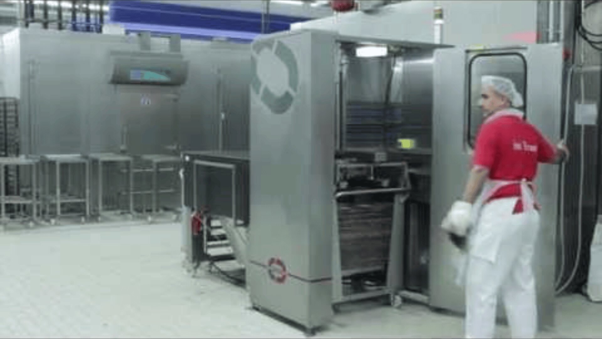 A worker in a red shirt, white pants, apron, and hairnet stands near industrial kitchen equipment, operating a large stainless steel machine in a clean, modern food processing facility.